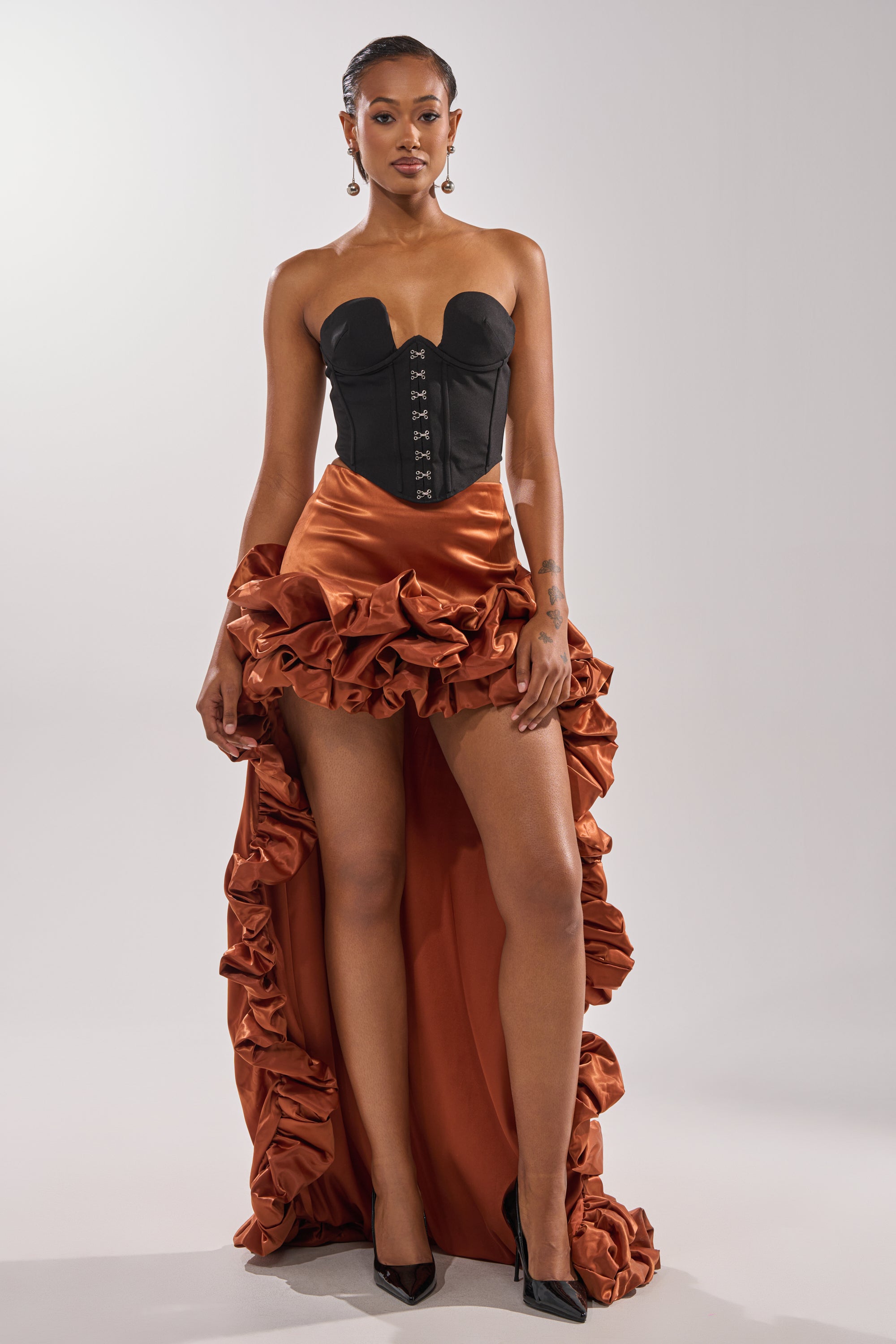 A woman stands confidently in the LOOK AT ME GO SATIN HIGH LOW SKIRT with orange hues, paired with a strapless black corset top and black heels—creating an eye-catching look perfect for any special occasion.