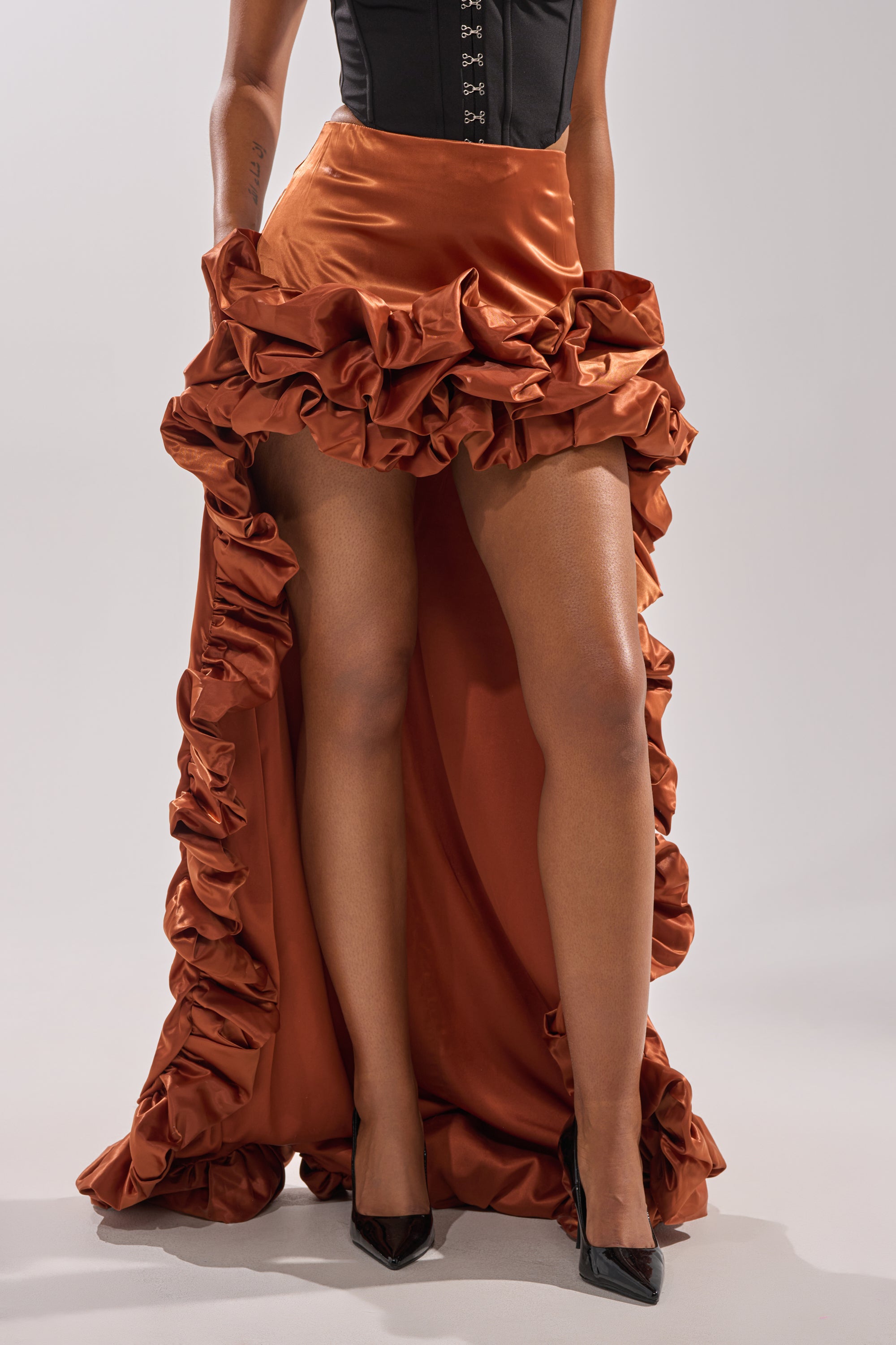 A person wearing a black corset top, the LOOK AT ME GO SATIN HIGH LOW SKIRT in vibrant orange, and black pointed heels, standing against a plain light background.