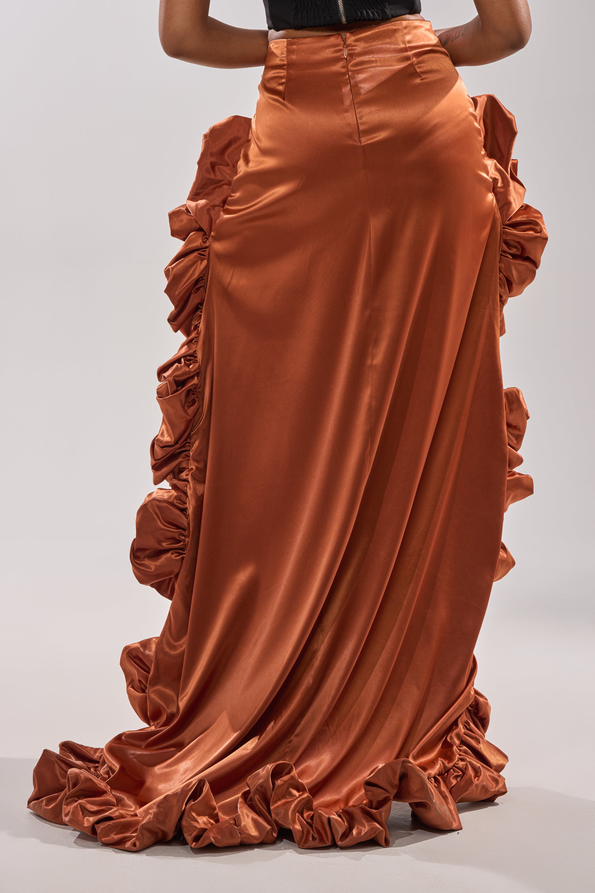 A woman models the LOOK AT ME GO SATIN HIGH LOW SKIRT in rust-orange, featuring striking ruffles and intricate side detailing, shown from the back against a plain light background—ideal for special occasions.