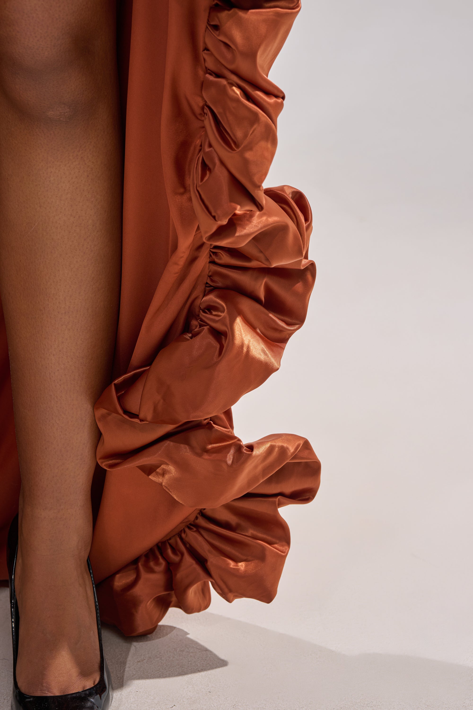 A close-up of a person wearing the LOOK AT ME GO SATIN HIGH LOW SKIRT in rust with dramatic side ruffles, paired with black heels; only one leg is visible against a plain background, ideal for special occasions.