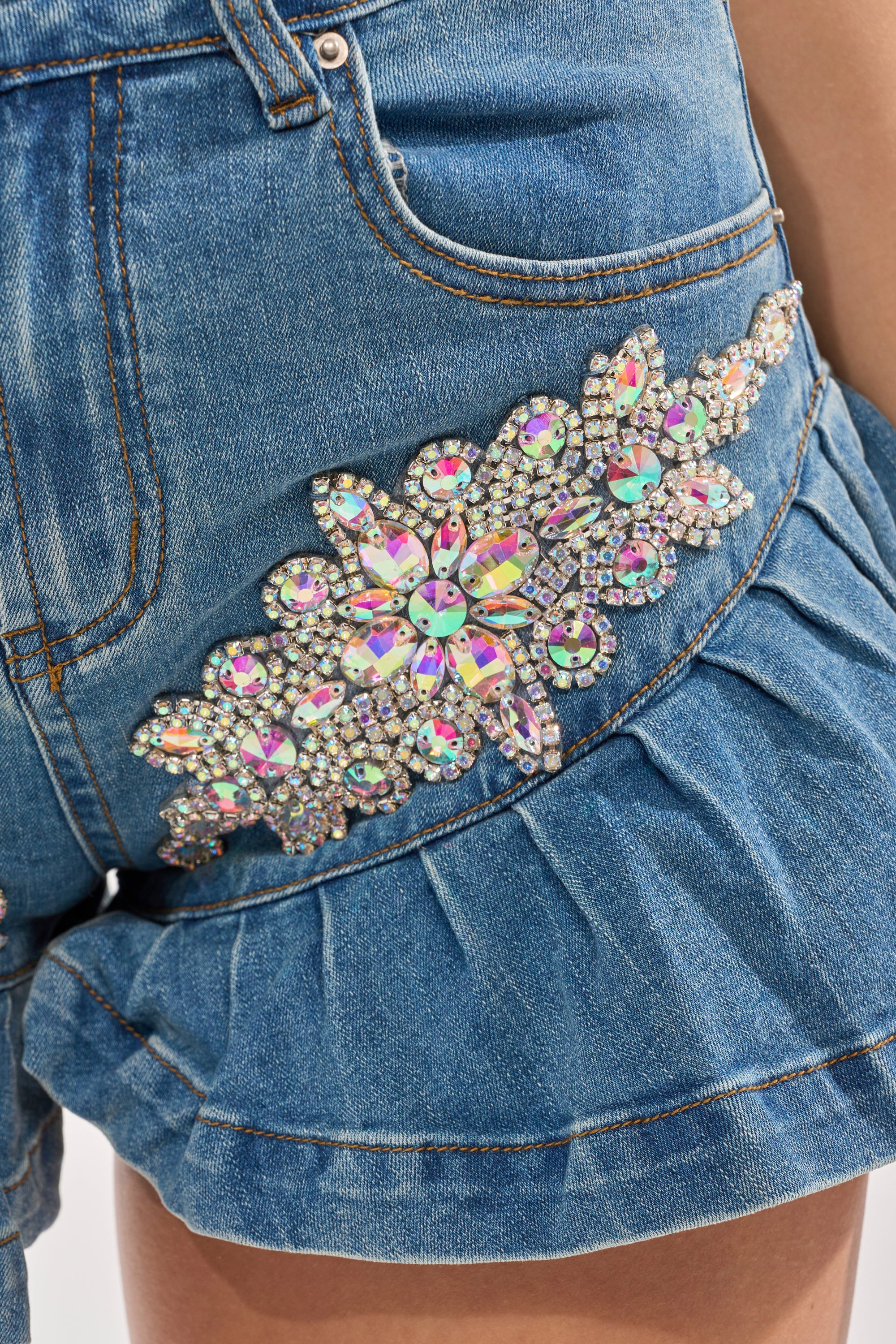 Close-up of the PRETTY GIRL PROBLEMS DENIM SHORT: Y2K-inspired pleated denim mini shorts with a ruffled hem and a large iridescent rhinestone flower embellishment near the pocket. 7