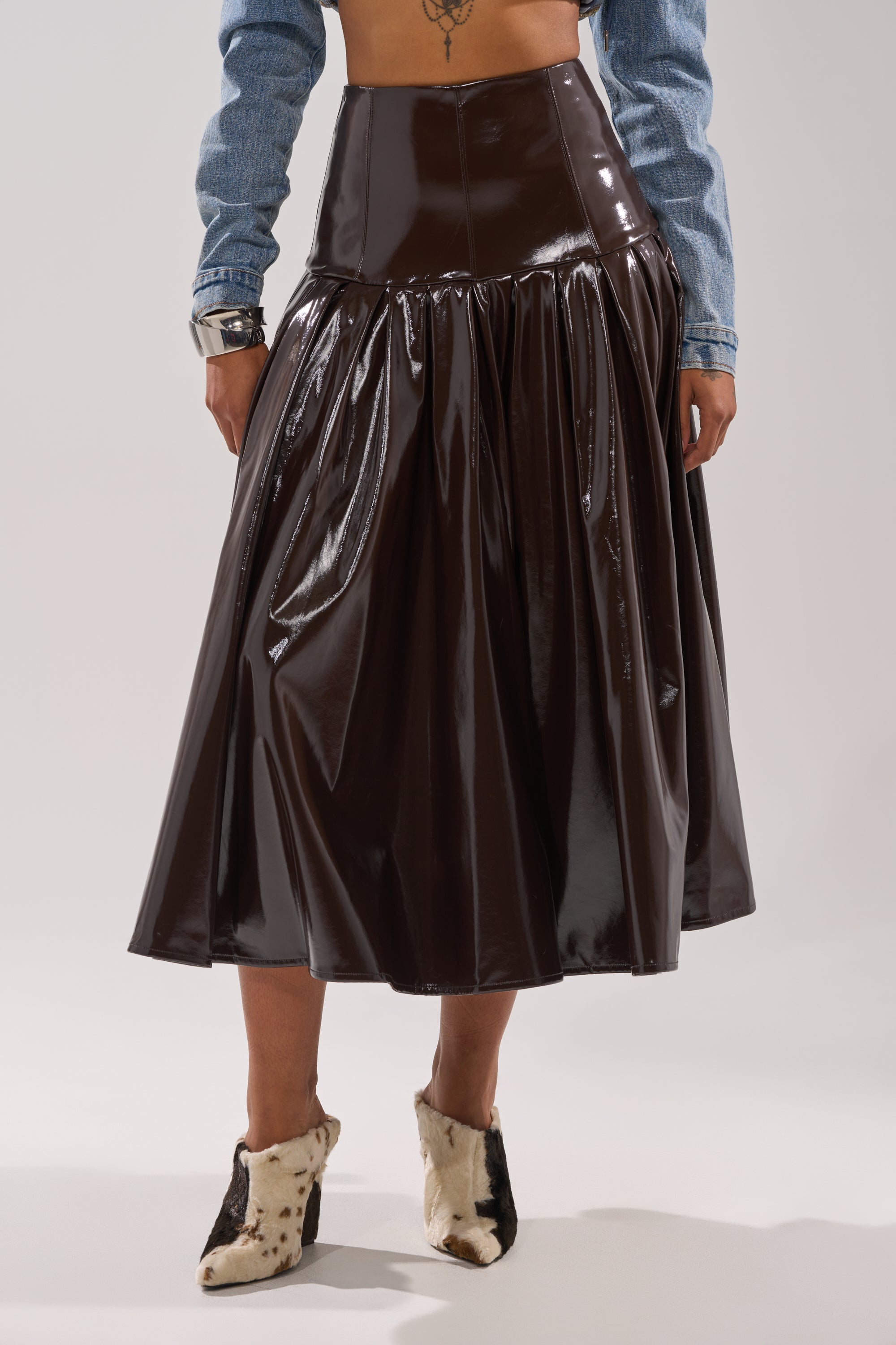 Wearing the BABY GIRL FAUX LEATHER MAXI SKIRT in shiny dark brown with a cropped denim jacket and cow-print ankle boots, this person stands against a plain light background, showing their midriff and tattoo. 1