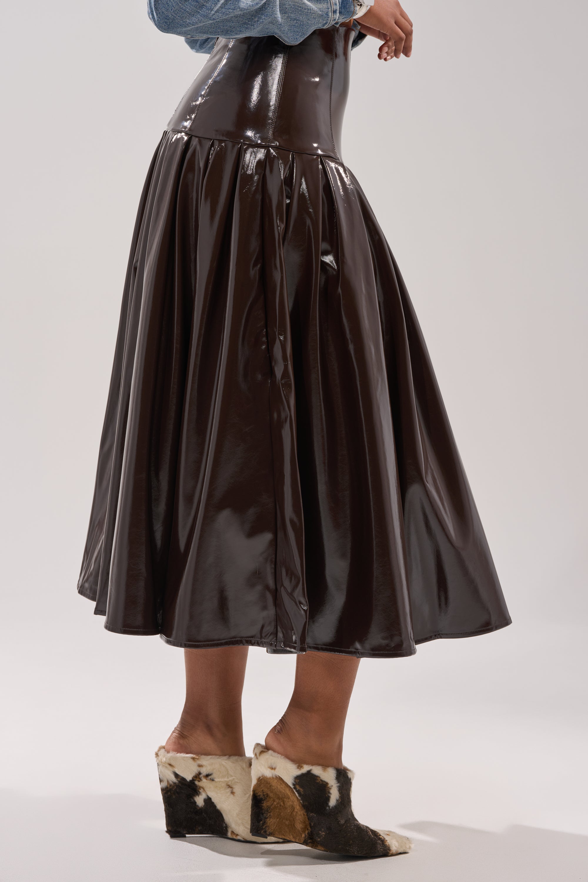 A person wears the BABY GIRL FAUX LEATHER MAXI SKIRT in shiny brown with a light blue denim top and black, white, and brown cowhide slip-ons. The photo is cropped at the shoulders. 2