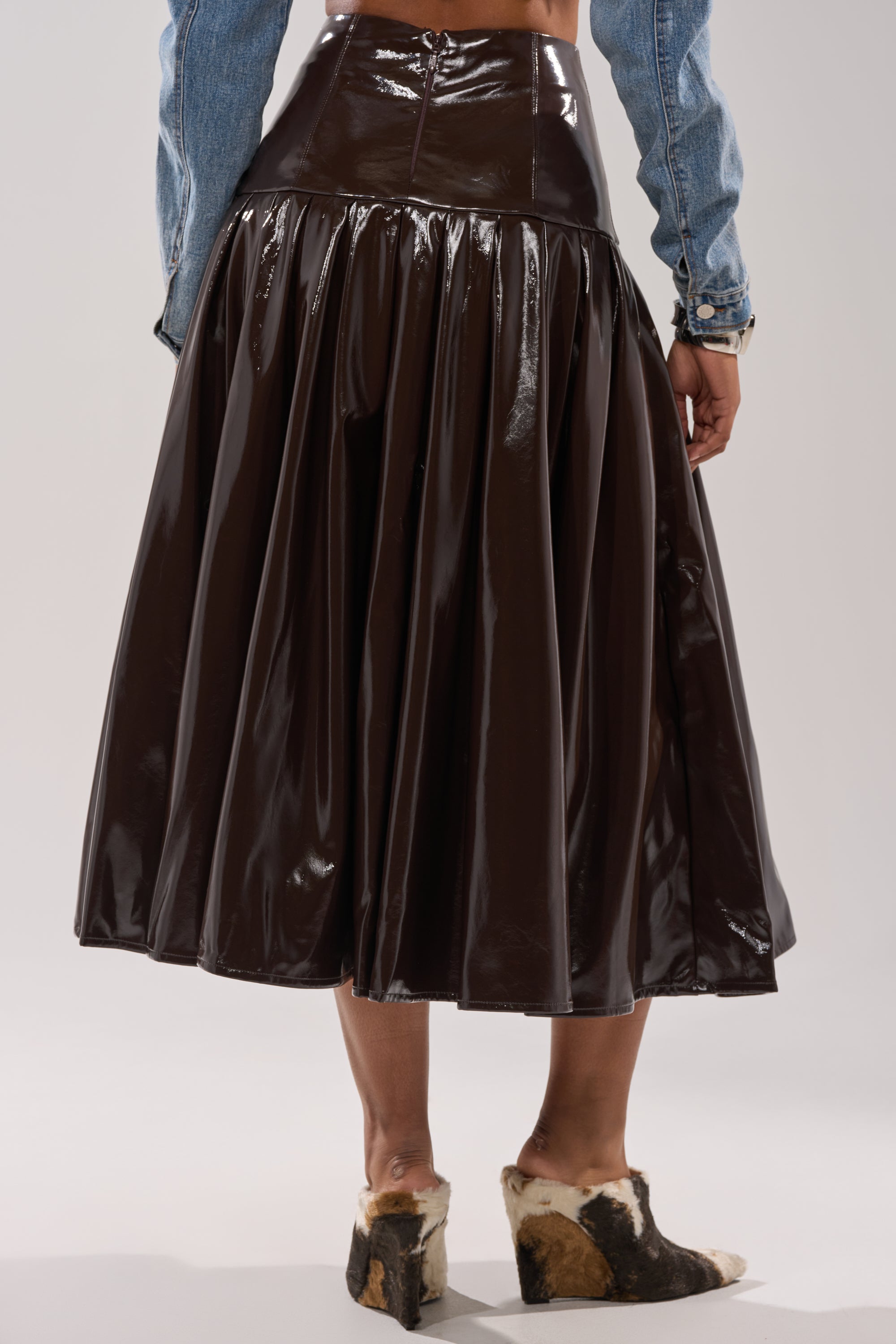 A person wears the BABY GIRL FAUX LEATHER MAXI SKIRT, a shiny dark brown high-waisted pleated ruffle skirt, styled with a light blue denim jacket and cowhide-patterned slip-ons. The photo shows the outfit from behind. 3