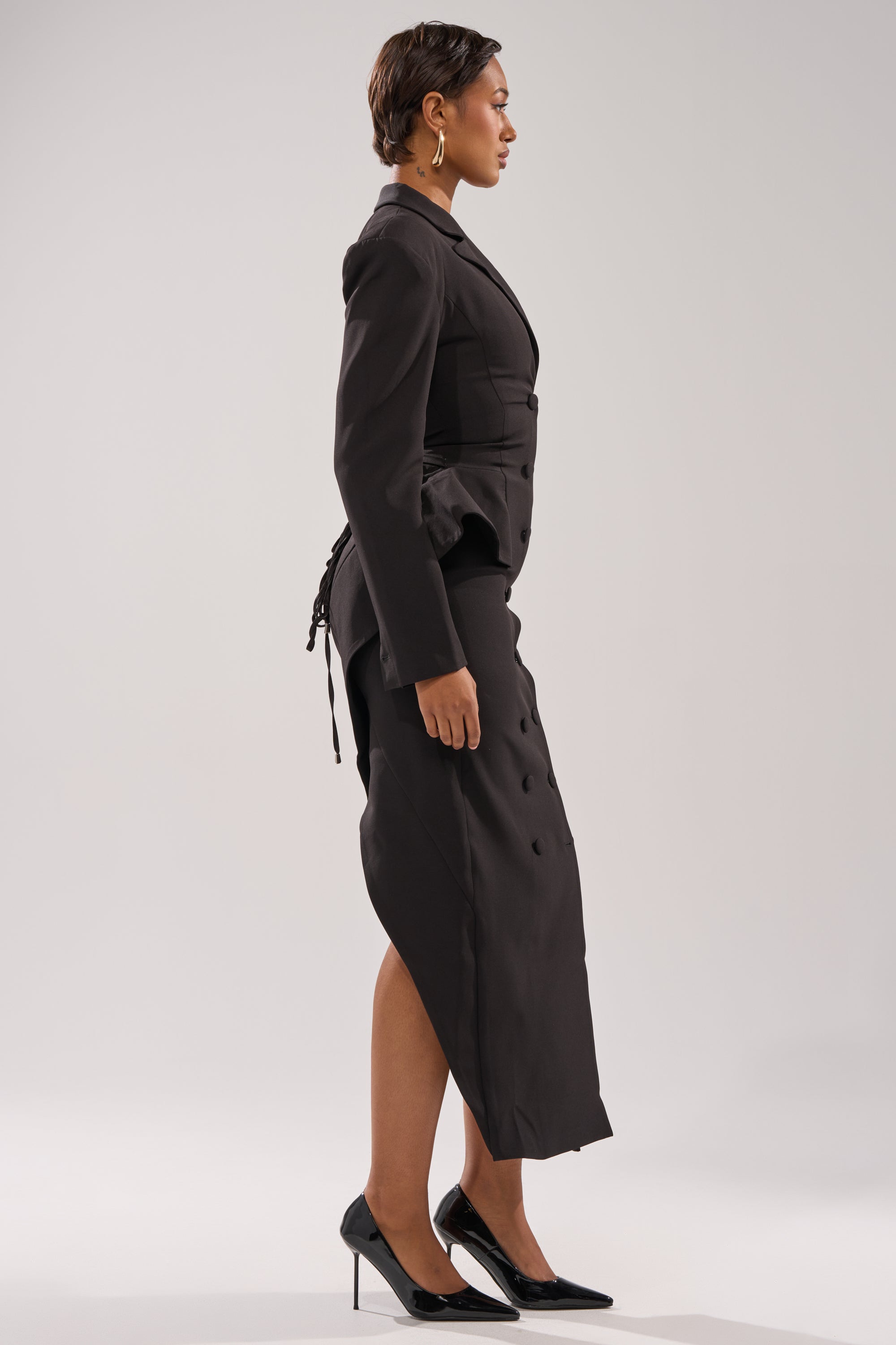 A woman stands in profile against a plain background, wearing the KIMMY BLAZER MAXI DRESS. She has short hair, hoop earrings, and black high-heeled shoes.