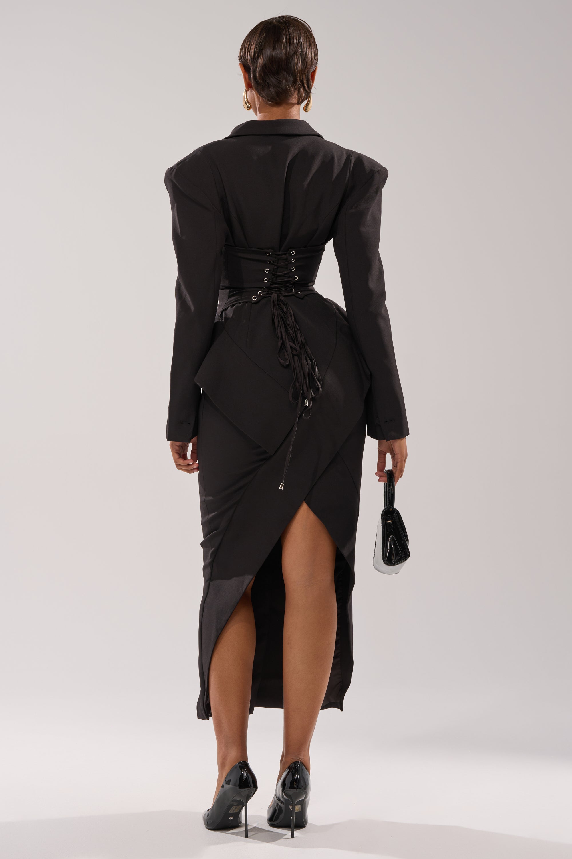 A person stands facing away, wearing the KIMMY BLAZER MAXI DRESS in black suiting fabric with structured shoulders, a corset-style laced waist, and a high-low hem. They hold a small black handbag and wear black heels.