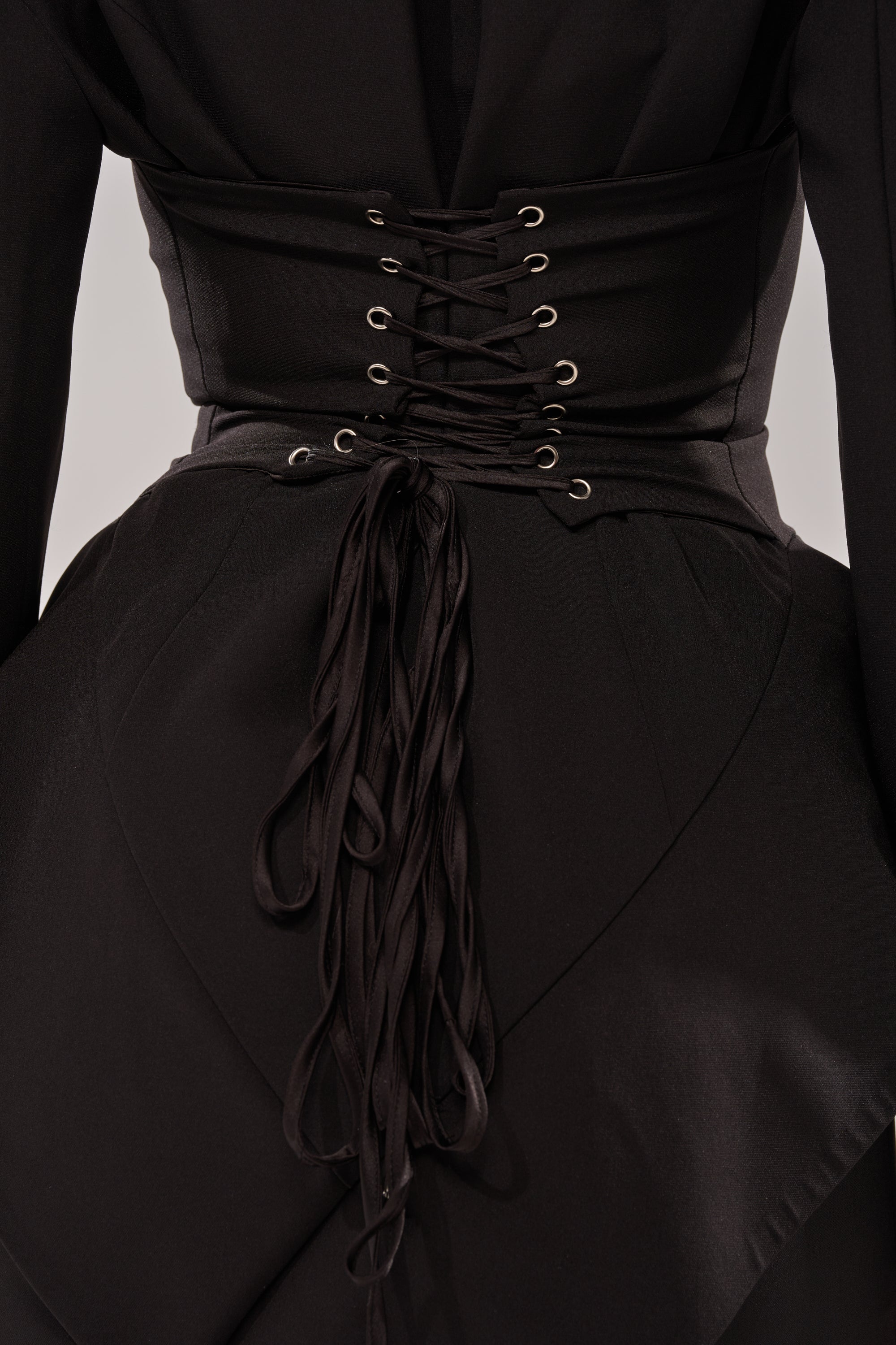 A close-up of a person in the KIMMY BLAZER MAXI DRESS, featuring a wide corset-style belt with crisscross laces and tied cords at the back. The structured black fabric creates an elegant office siren look.