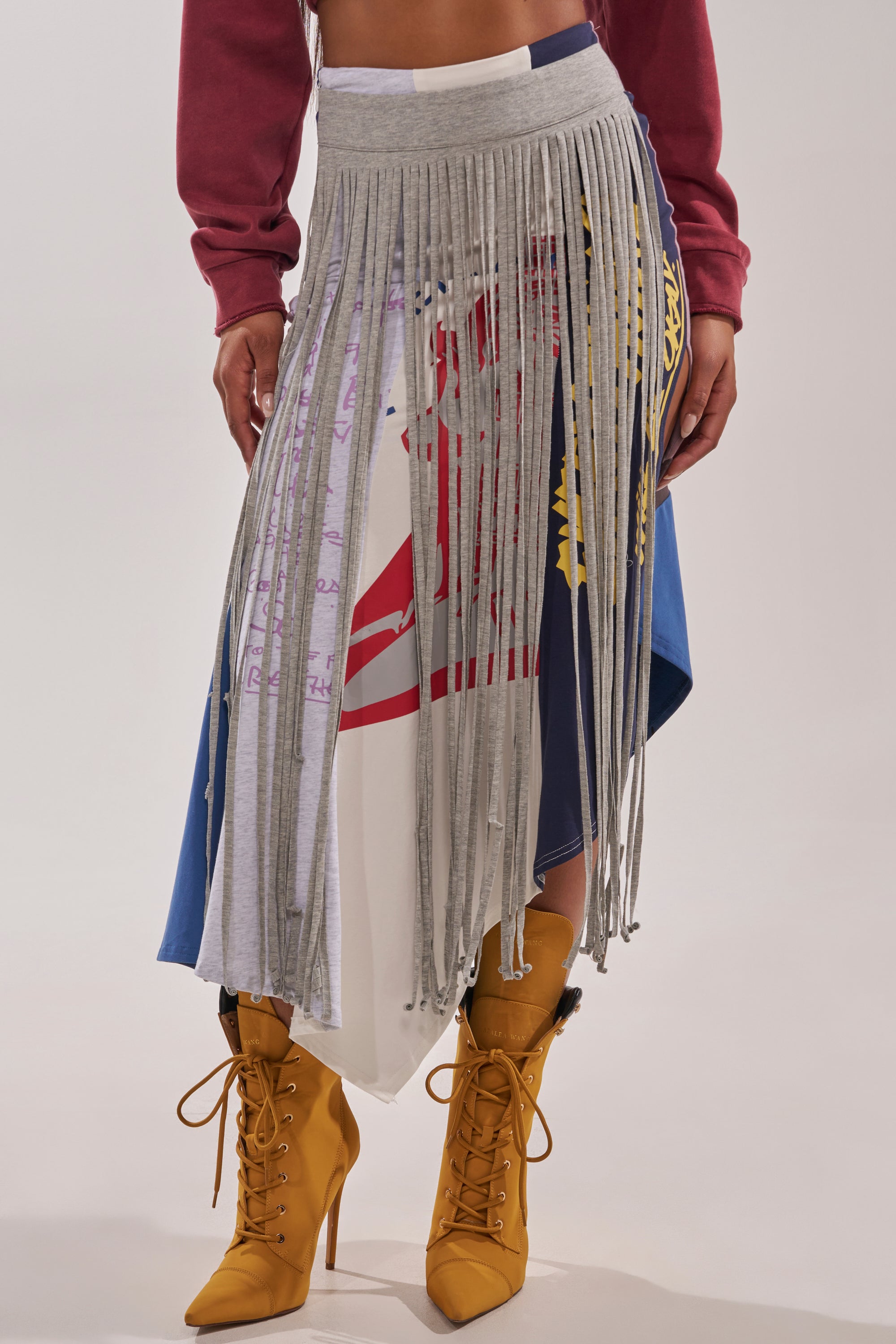 A person wears the STRESSED AND DISTRESSED MIDI SKIRT, featuring gray fringe patchwork and graphic prints over blue and white asymmetrical fabric, paired with mustard yellow lace-up heeled boots. Only their hands and midsection are partly visible. 0