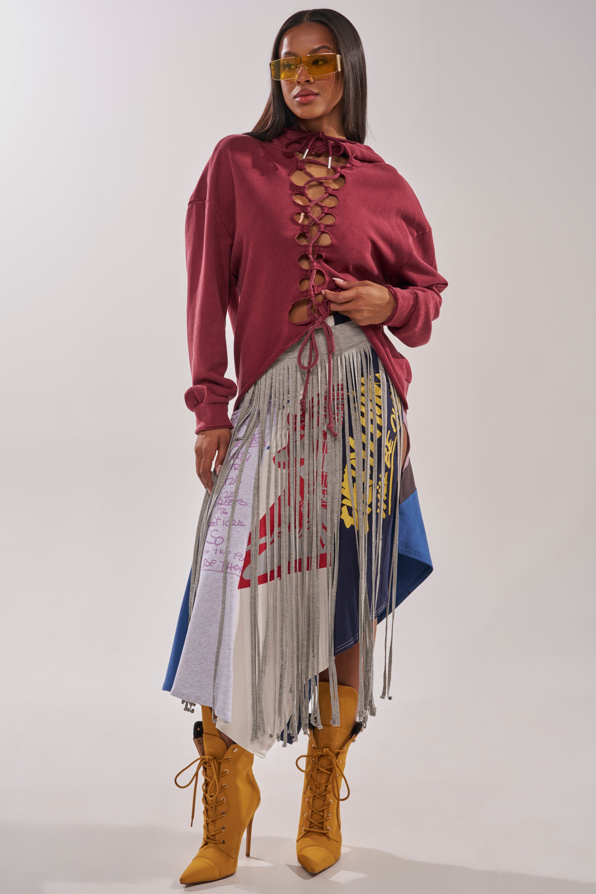 A woman poses confidently against a plain background in yellow sunglasses, a maroon lace-up top, and the STRESSED AND DISTRESSED MIDI SKIRT—a multicolored fringe patchwork midi skirt with graphic designs—paired with mustard yellow heeled boots. 8