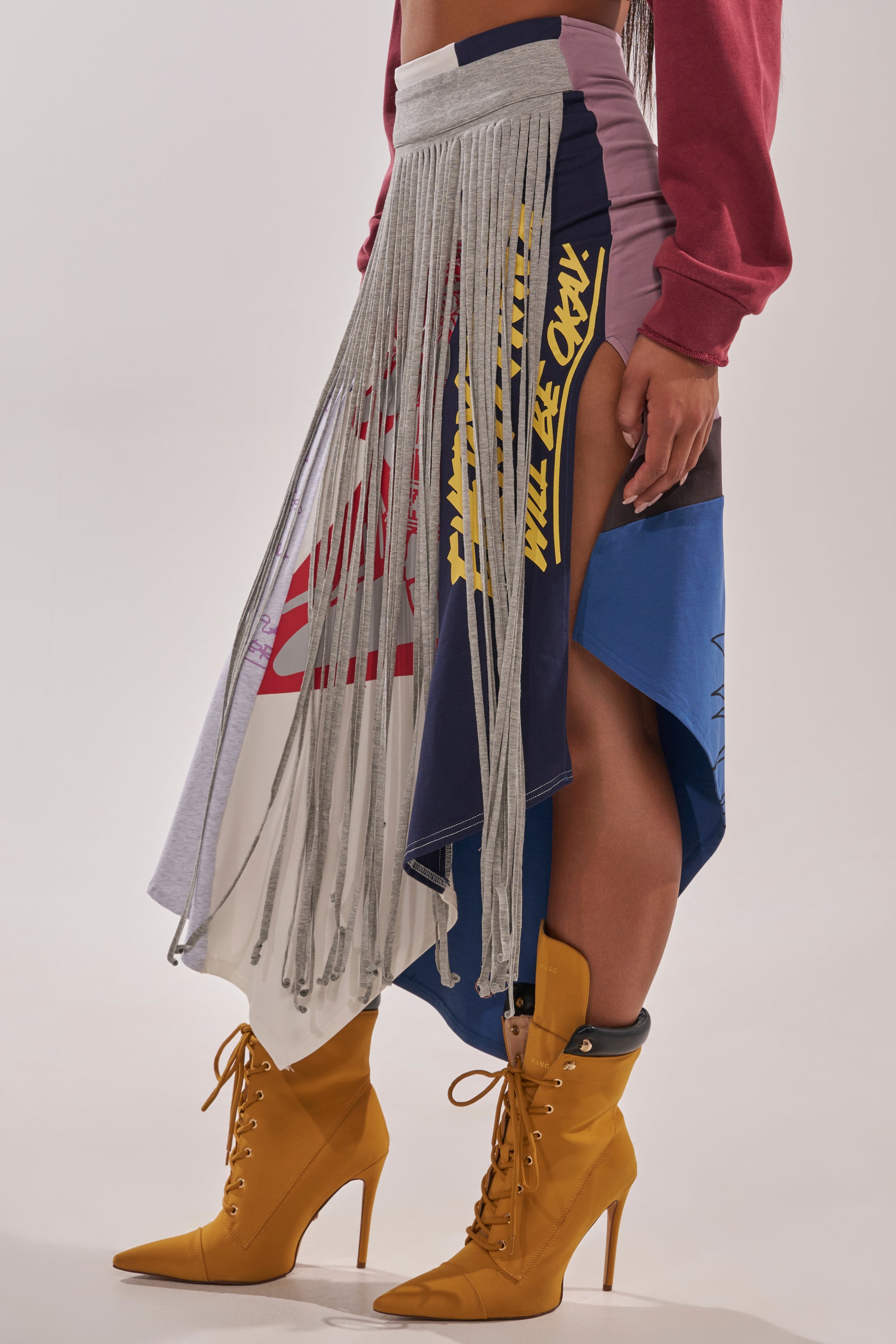 A person wears the STRESSED AND DISTRESSED MIDI SKIRT—a multicolored, deconstructed piece with long gray fringe, high slit, and text accents—paired with mustard yellow heels and a red sweatshirt. 2