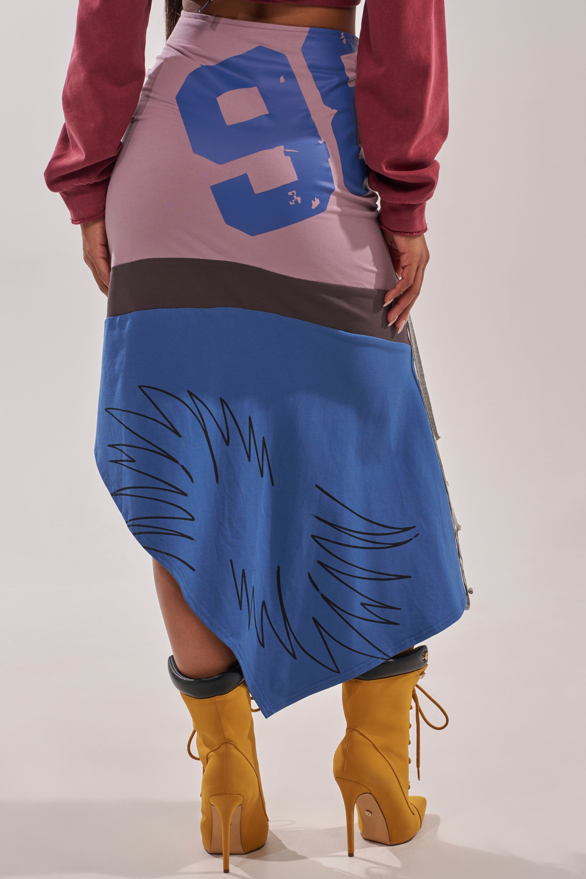A person stands with their back to the camera, wearing the STRESSED AND DISTRESSED MIDI SKIRT with numbers and graphic designs, paired with a red long-sleeve top and yellow lace-up heeled boots. 4