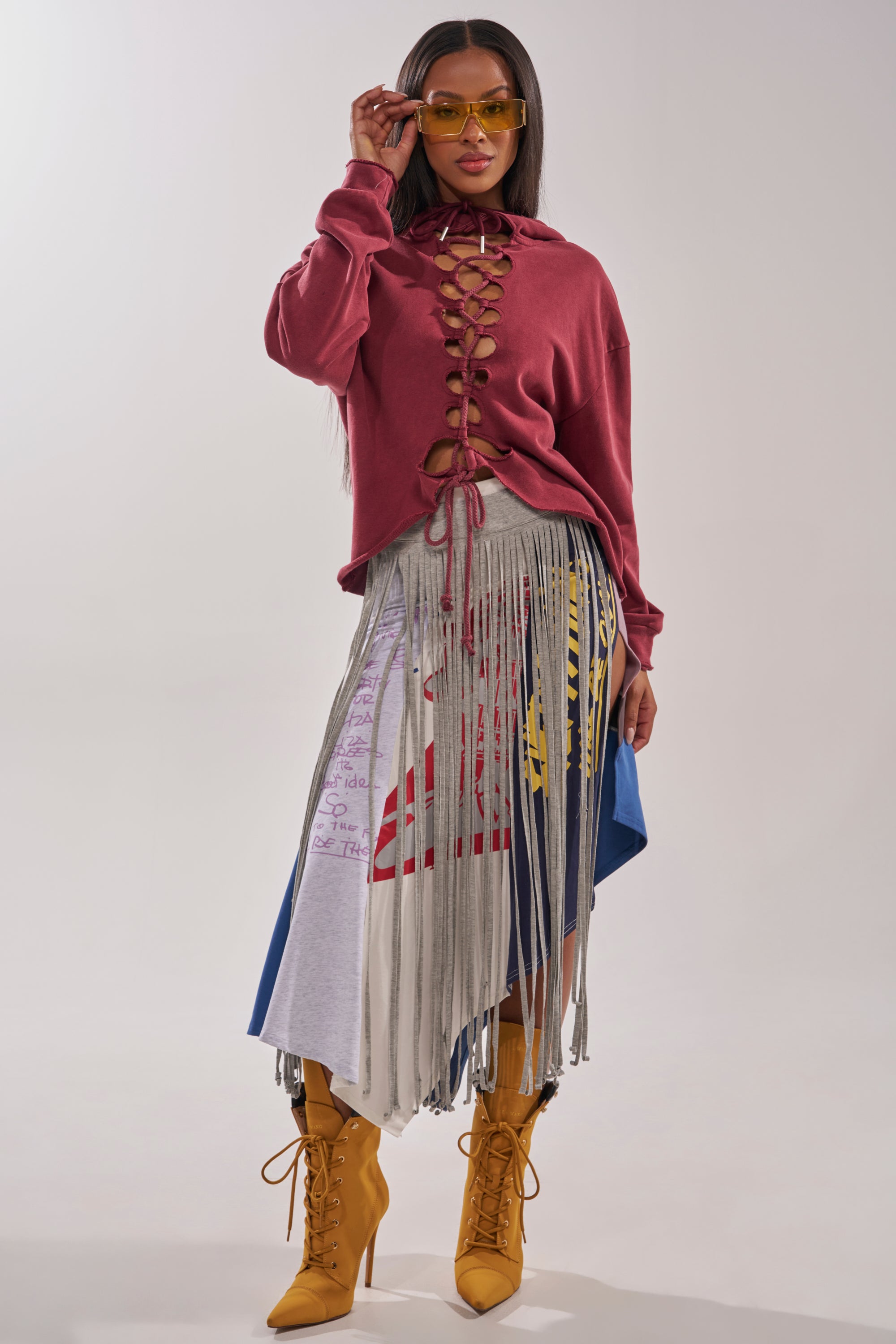 A woman poses in a burgundy lace-up sweatshirt, the STRESSED AND DISTRESSED MIDI SKIRT with fringe and graphic prints, yellow heeled boots, and yellow sunglasses against a plain light background. 5