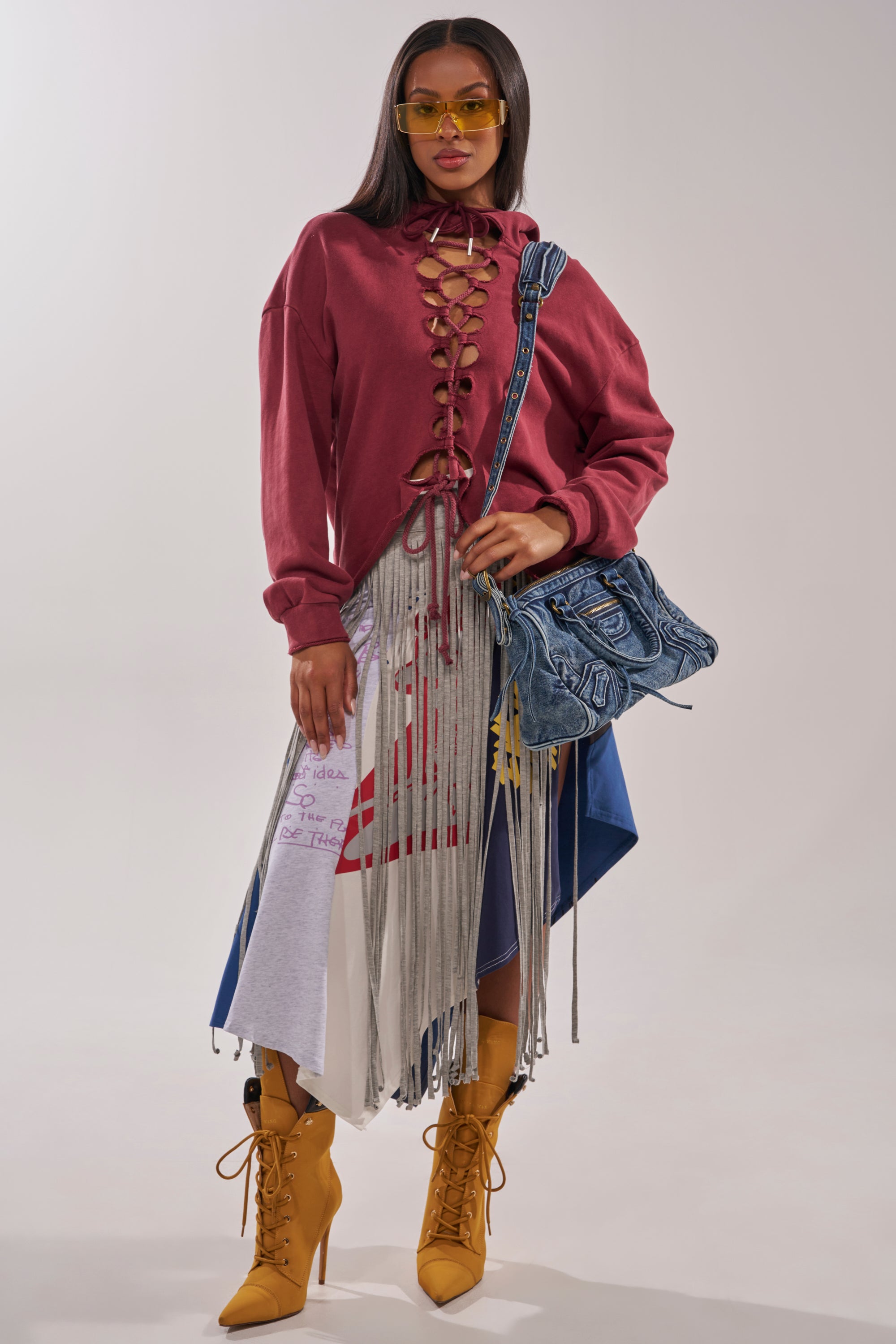 A woman stands confidently in yellow sunglasses, a maroon lace-up top, yellow boots, and the STRESSED AND DISTRESSED MIDI SKIRT with silver fringe and graphic designs, carrying a large blue denim bag over her shoulder. 6