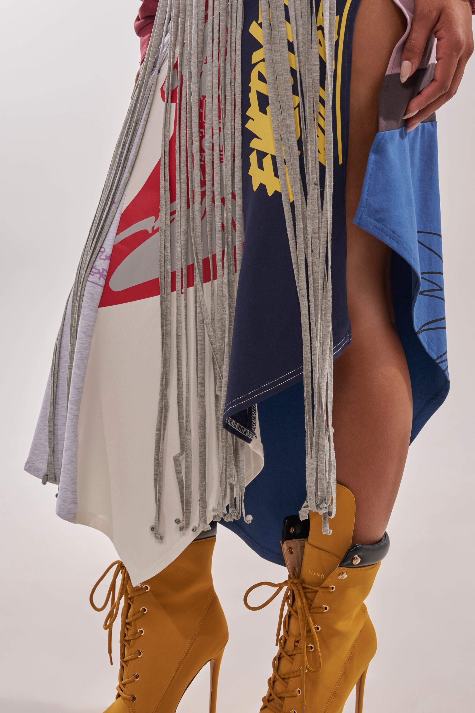 A person models the STRESSED AND DISTRESSED MIDI SKIRT—a multicolored fringe patchwork design with graphic prints—paired with mustard yellow high-heeled lace-up boots. Only their lower half is visible against a plain background. 7
