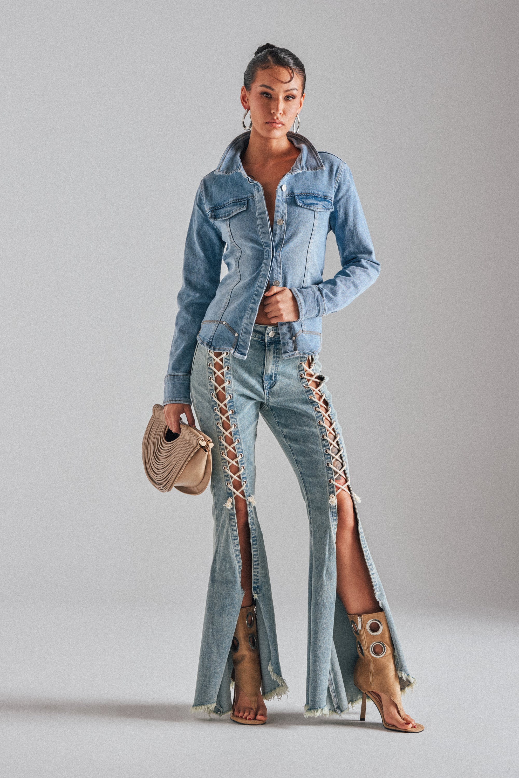 A woman faces forward in the CRAZY ENOUGH LACE UP FLARE DENIM, paired with a denim jacket, tan high-heeled boots, and a beige clutch. The pants have front slits and lace-up detailing. She stands against a plain light gray background.