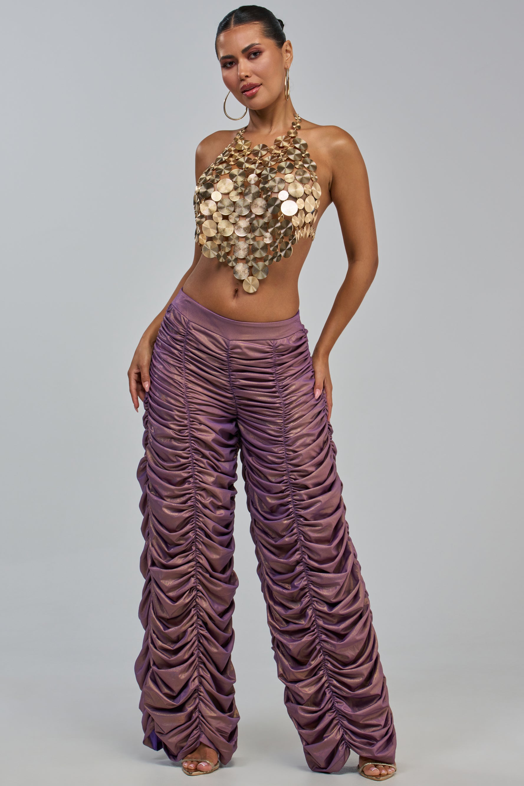 A woman poses in the WOW WOW WOW RUCHED PANT, a pair of high-waisted, ruched rose gold pants. She completes her look with a halter top crafted from large gold discs, hoop earrings, and strappy heels while standing against a plain gray background.