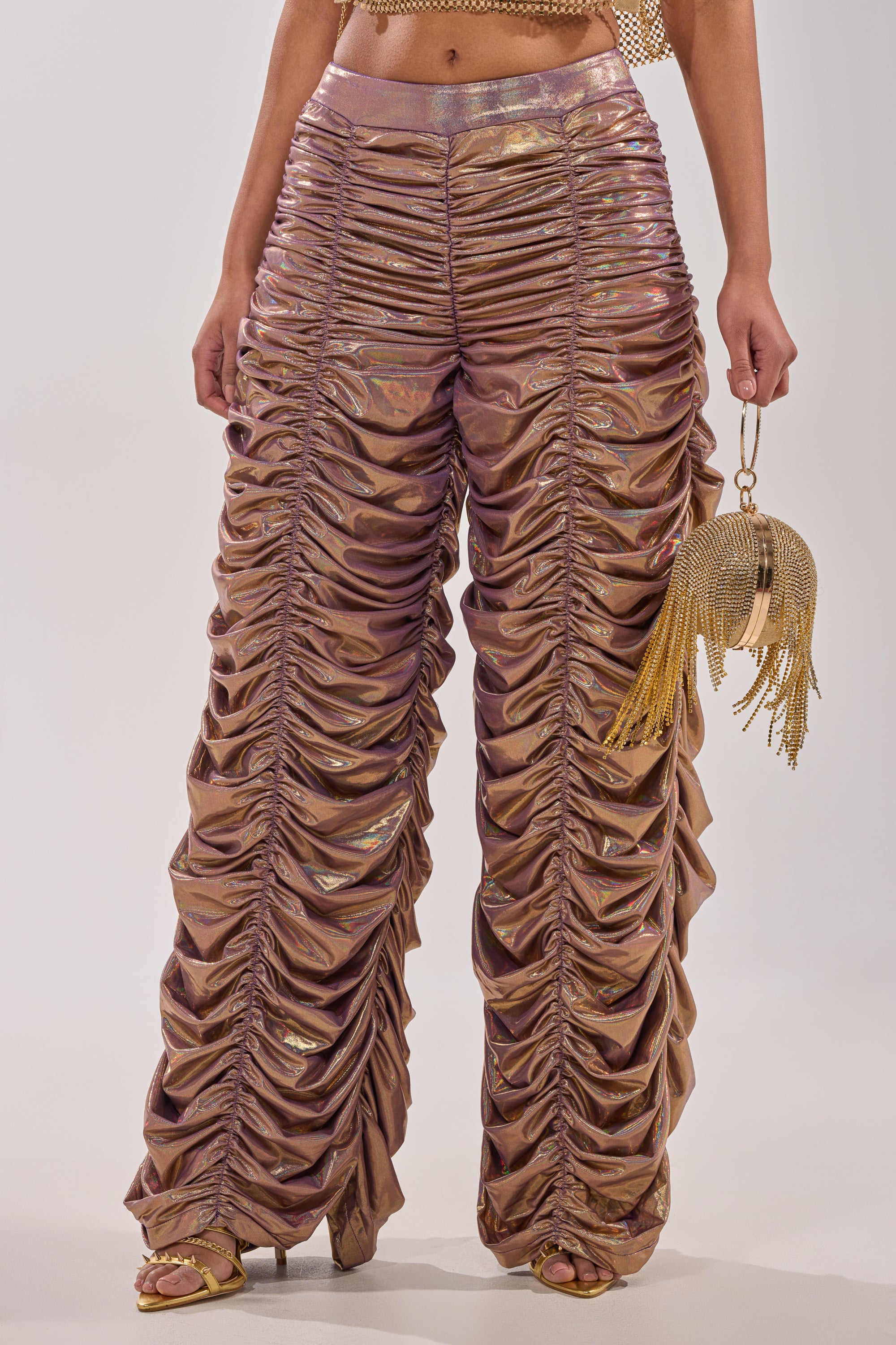 Wearing WOW WOW WOW RUCHED PANT in shiny rose gold, styled with gold open-toe shoes and a gold beaded fringe handbag. Photo shows the look from shoulders down against a plain background.
