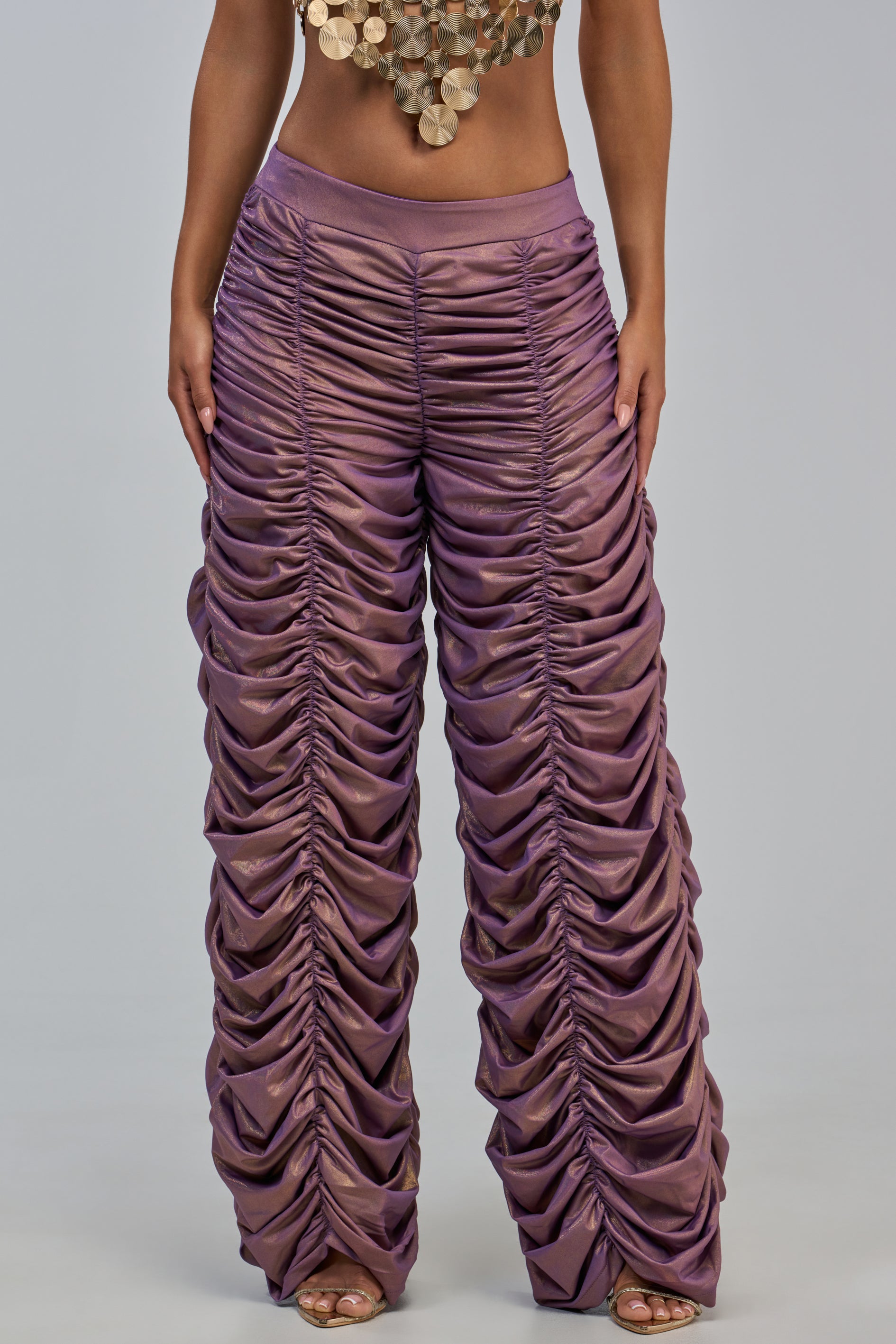 A person wearing the WOW WOW WOW RUCHED PANT in shiny rose gold, paired with a crop top featuring large gold discs, stands against a plain light background.
