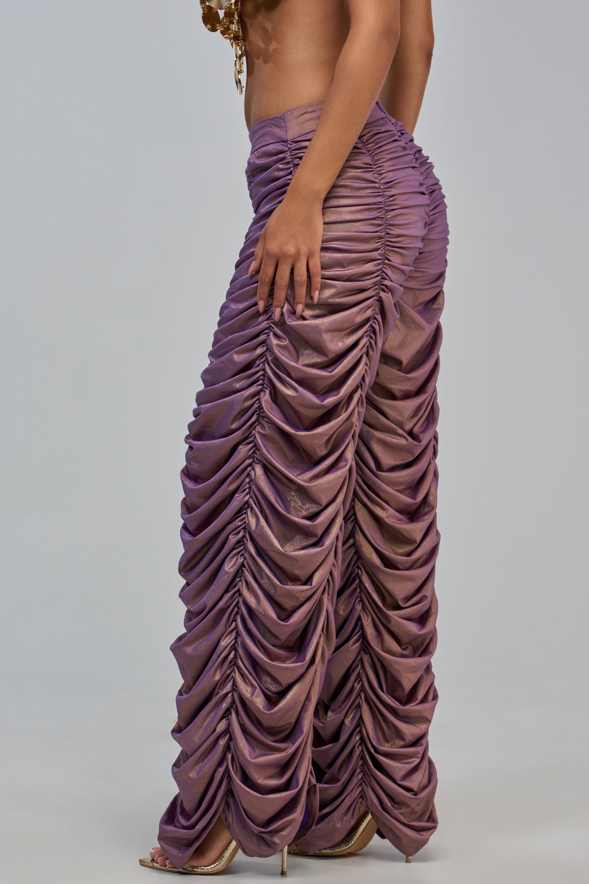 A person stands sideways against a plain light gray background, wearing the WOW WOW WOW RUCHED PANT—a statement, high-waisted lavender style featuring ruched fabric throughout for a textured, layered look reminiscent of metallic pants—paired with gold heels.