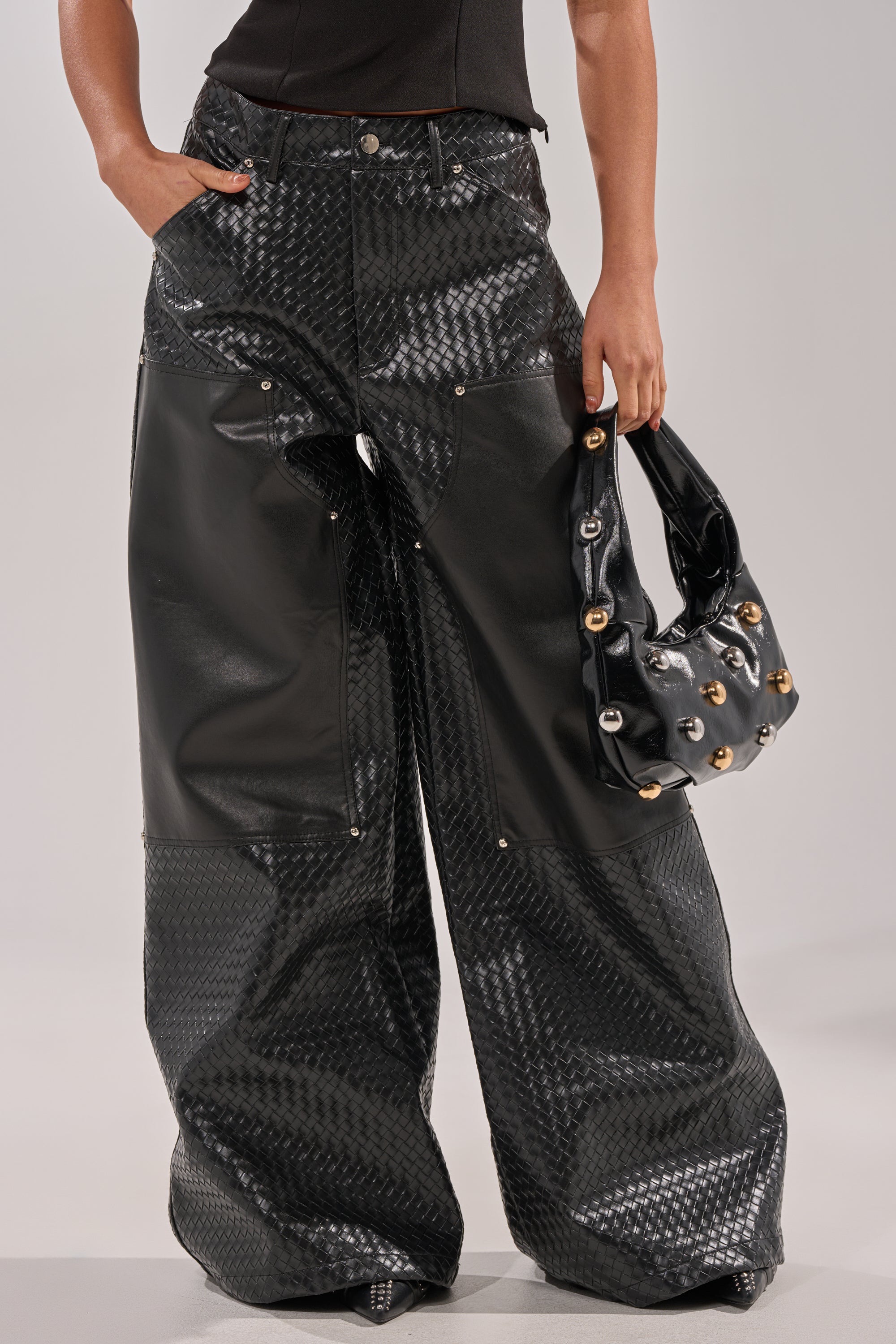 A person wears WILD TEXTURED FAUX LEATHER PANT—black, wide-leg street style pants with shiny panels—and carries a matching studded black handbag, standing against a plain light background. Upper body partially visible. 0