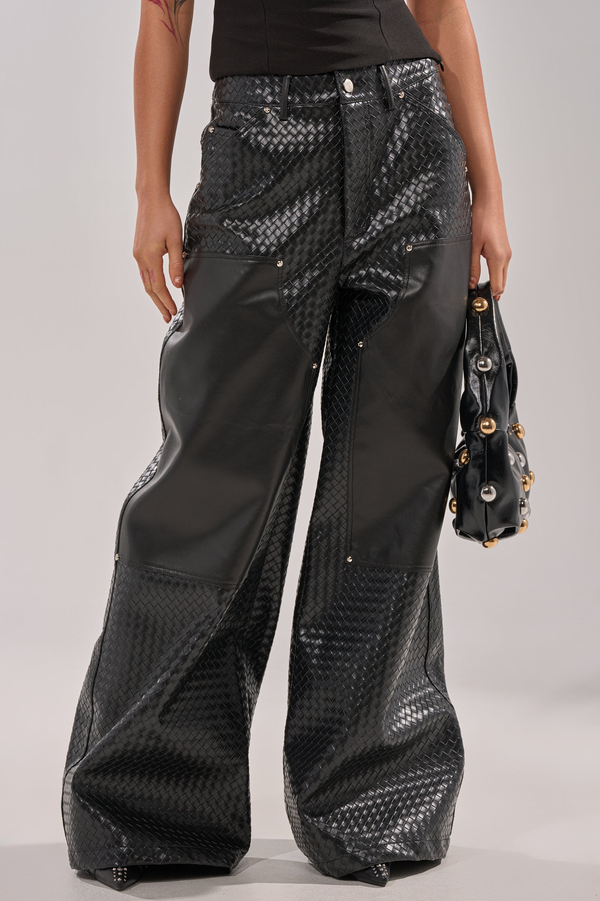 A person wearing WILD TEXTURED FAUX LEATHER PANT with wide-leg and patchwork details, holding a black handbag with gold accents, stands against a neutral background. 2