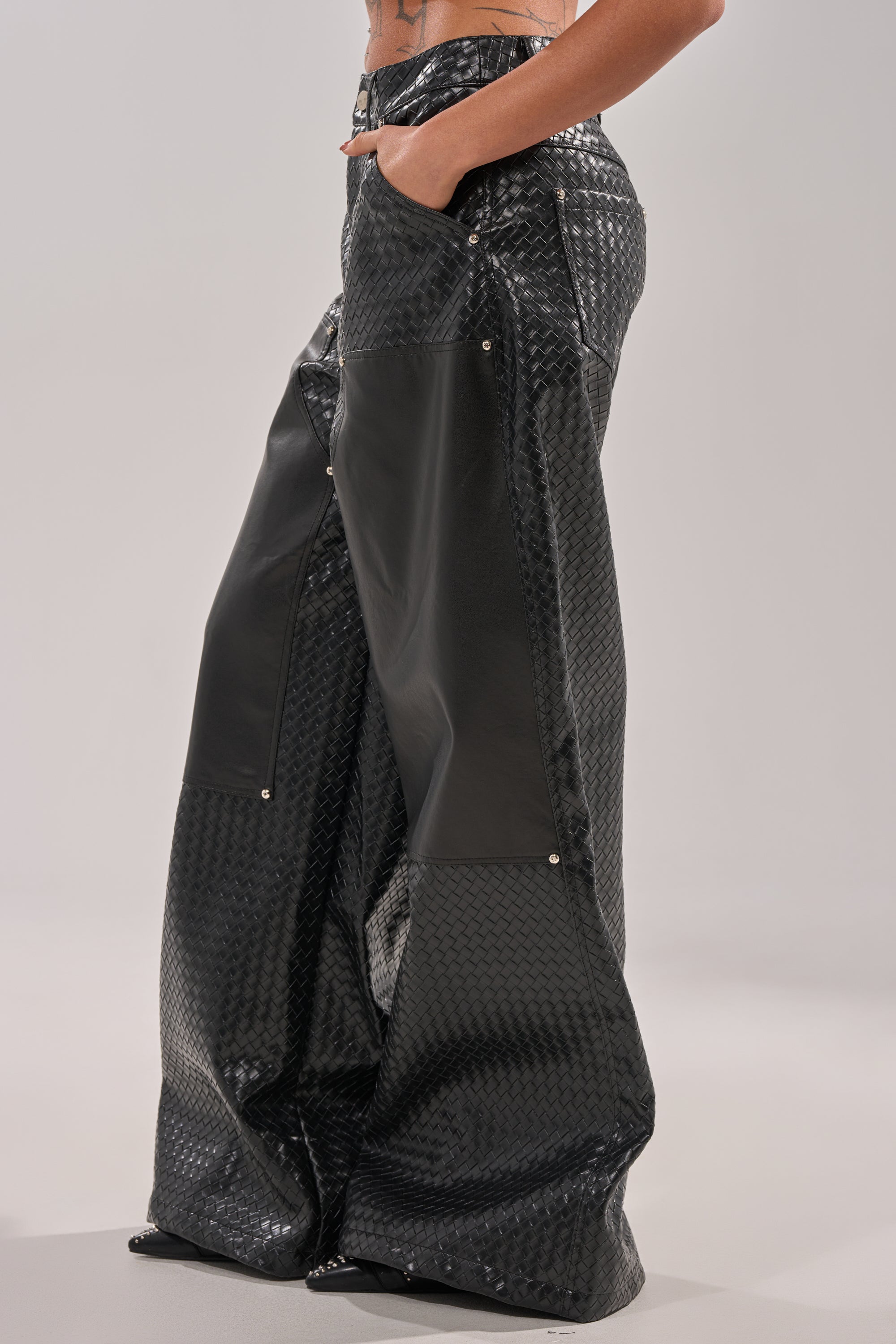 A person wears the WILD TEXTURED FAUX LEATHER PANT—high-waisted, wide-leg cargo pants with shiny textured faux leather and smooth black panels, featuring side pockets. Their hands are tucked in against a plain, light background. 3