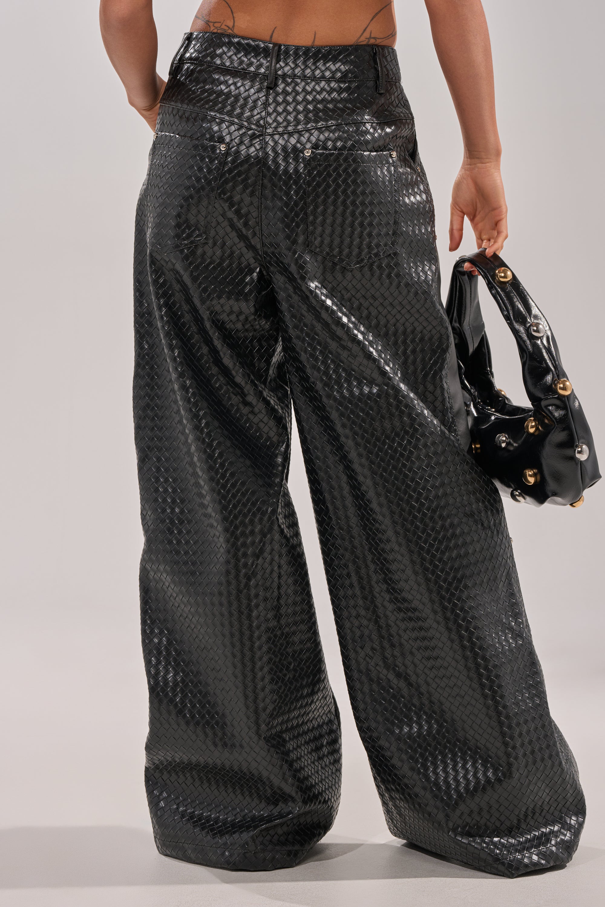 A person faces away, wearing WILD TEXTURED FAUX LEATHER PANT in a high-waisted, wide-leg fit with a textured look. In their right hand, they carry a black handbag accented with gold details. 4