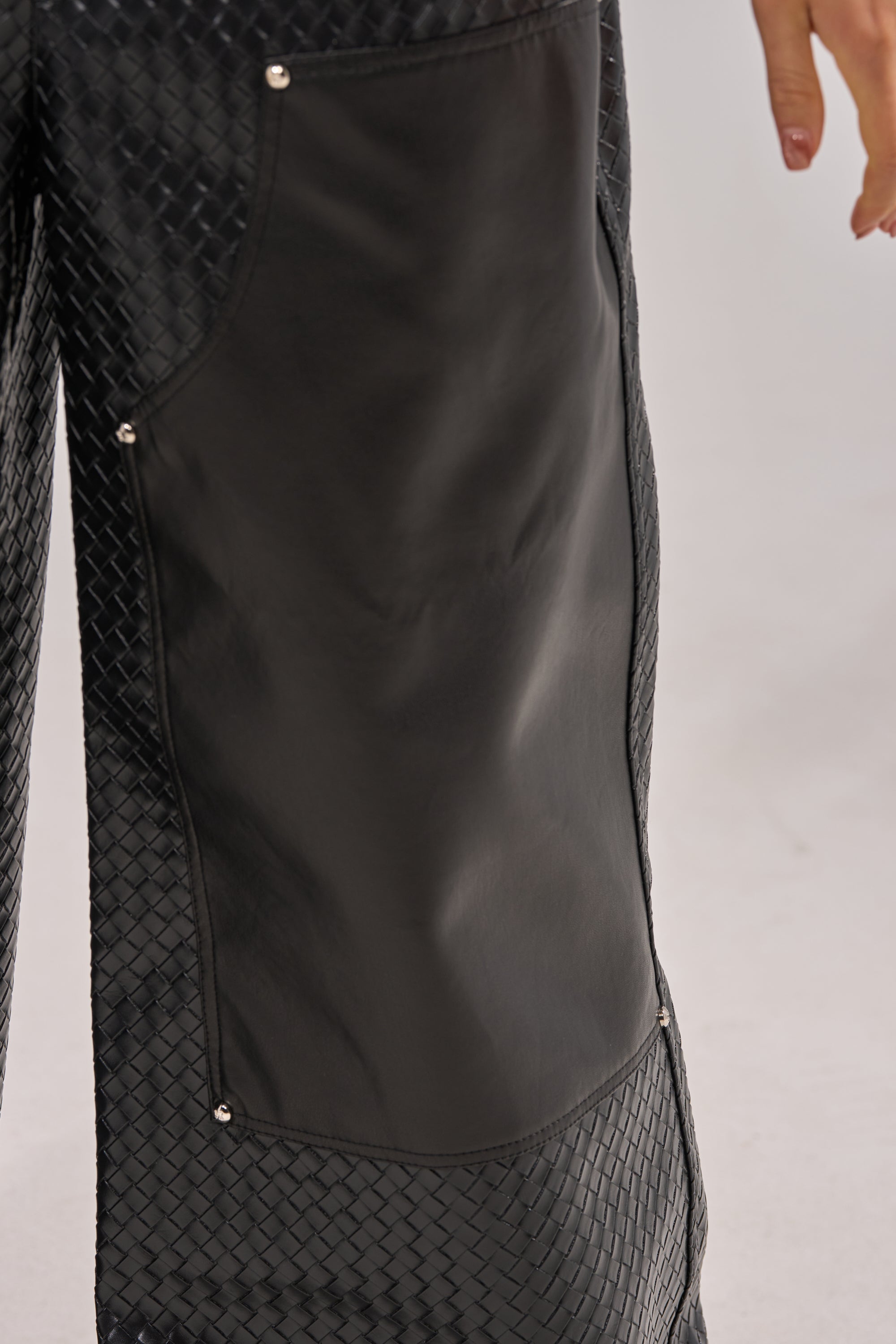 Close-up of a person wearing WILD TEXTURED FAUX LEATHER PANT, featuring wide legs, a large smooth leather patch, and metal rivets on the side. Part of a hand is visible on the right against a plain light background. 6