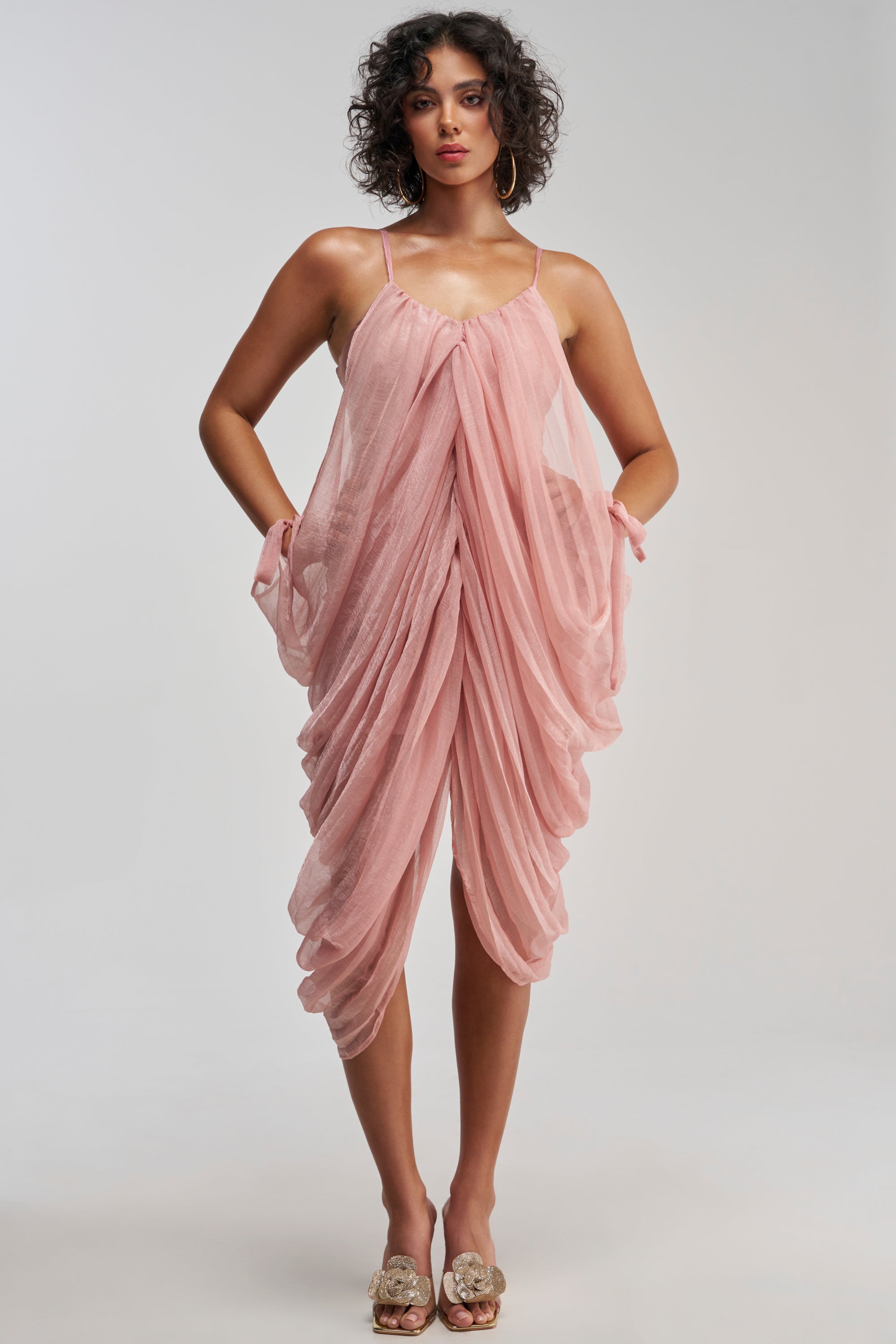 A woman in the VIVA LA VIDA MESH DRESS IN MAUVE, featuring a pinkish-mauve draped mini silhouette with thin straps, stands with her hands in the pockets against a plain backdrop. Her curly hair and open-toe shoes complete this modern homecoming-ready look.