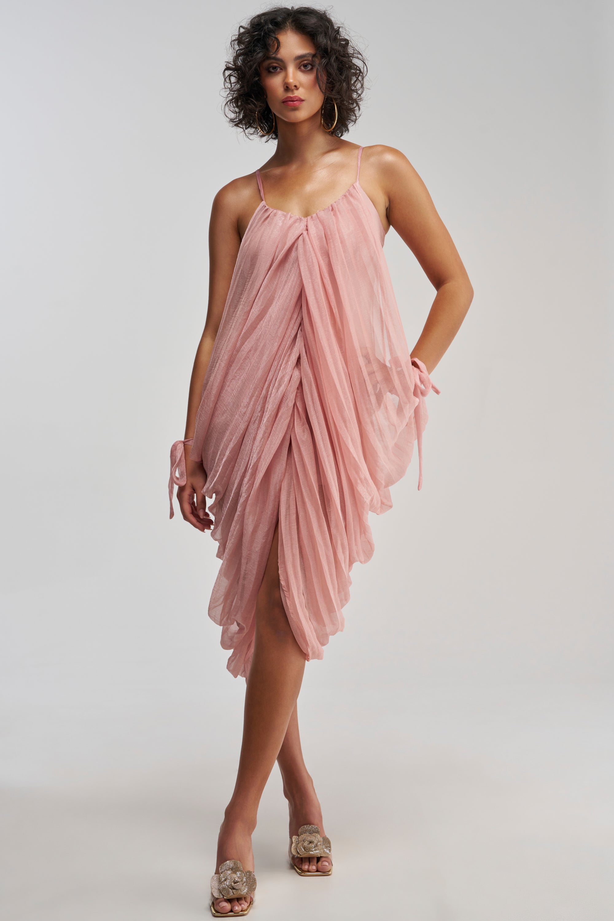 A woman wearing the VIVA LA VIDA MESH DRESS IN MAUVE, a soft pink, draped mini dress with spaghetti straps featuring a flowy, layered design and bow details at the wrists, paired with open-toe sandals. This elegant look is perfect as a homecoming dress and stands out beautifully against a plain background.