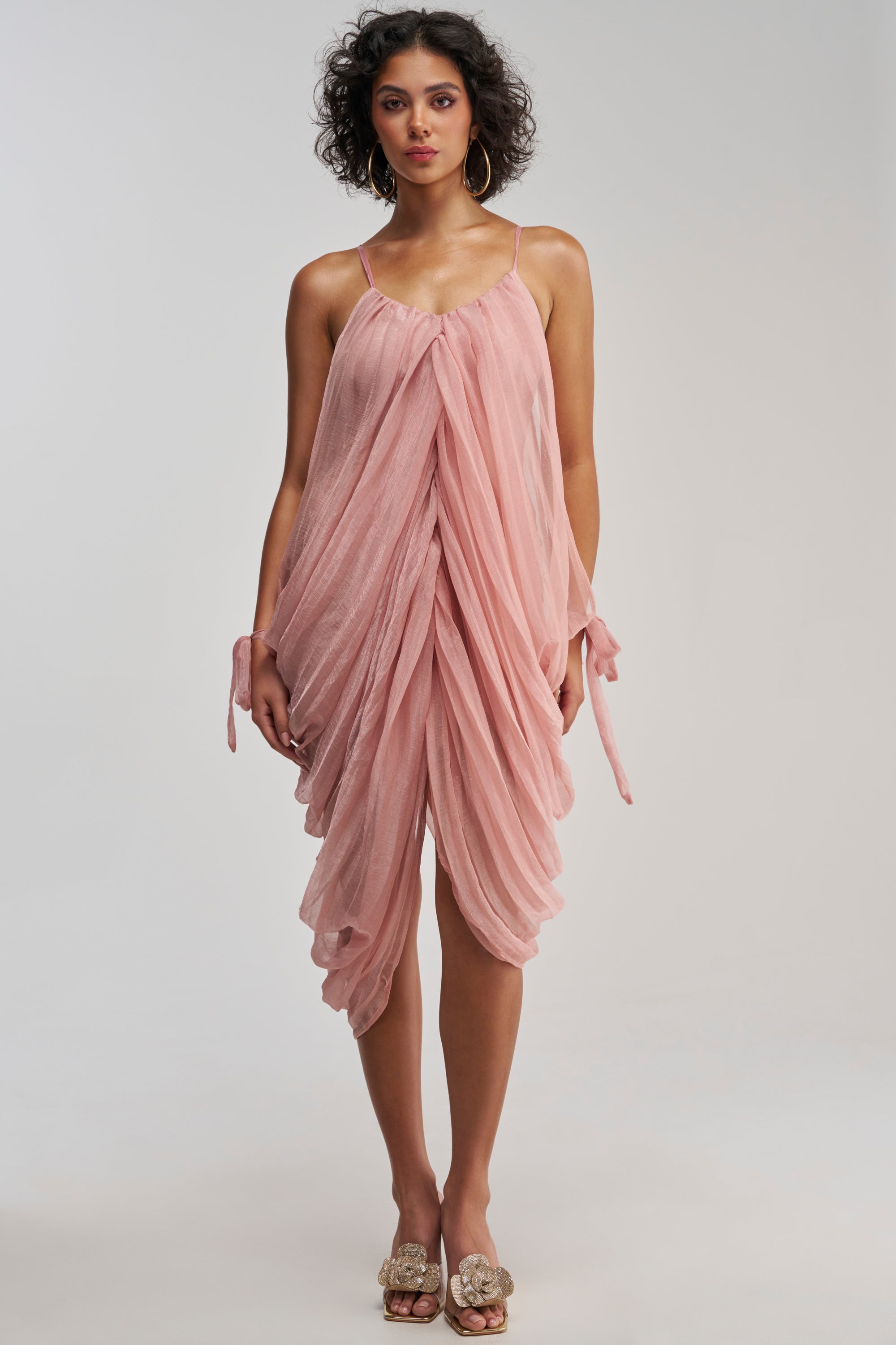 A woman stands facing forward, wearing the VIVA LA VIDA MESH DRESS IN MAUVE—a sleeveless mini dress with thin straps and draped, ruffled mesh layers. She has curly hair, hoop earrings, and slip-on sandals with decorative bows.