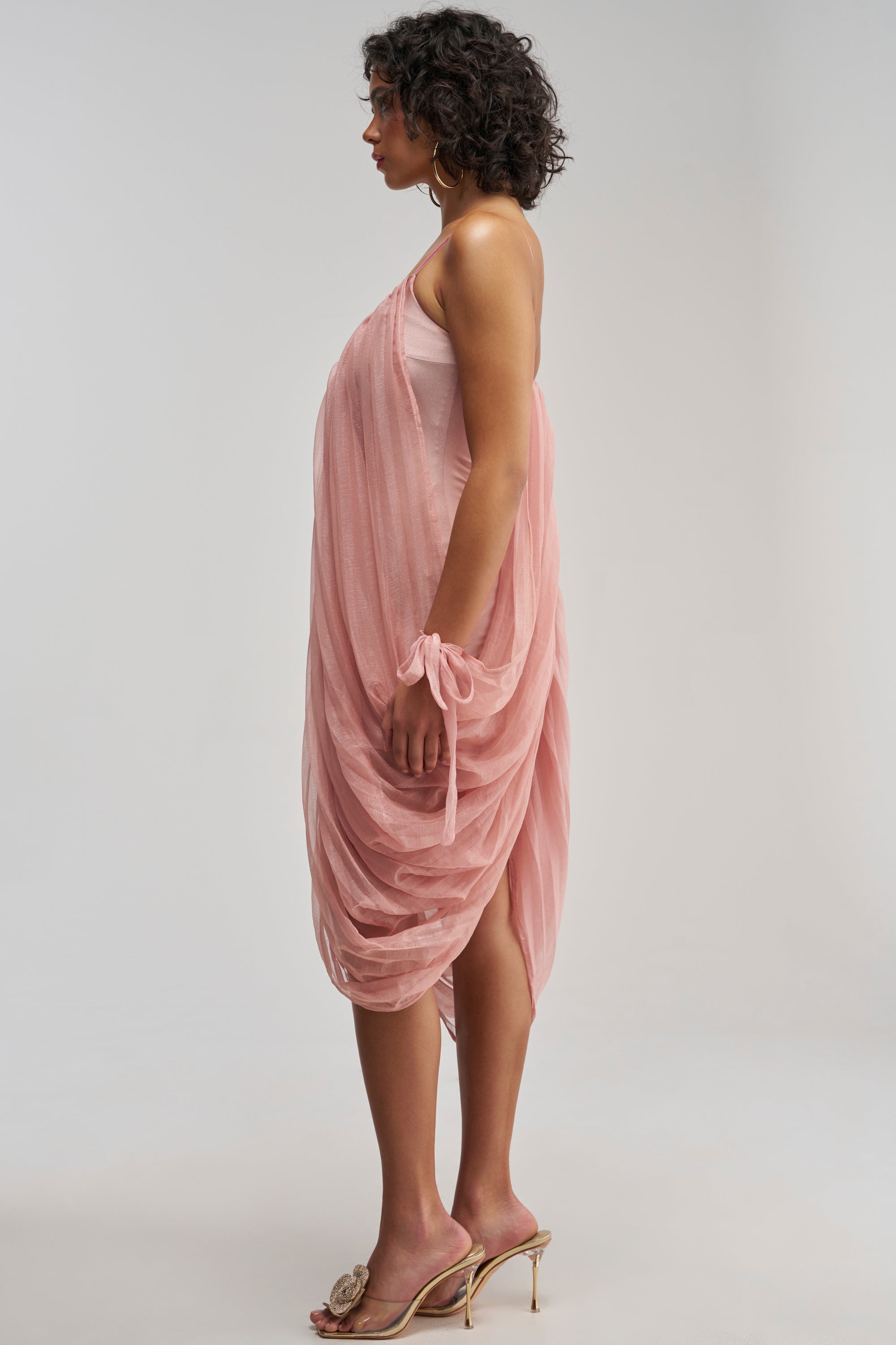 A woman stands in profile against a plain light background, wearing the VIVA LA VIDA MESH DRESS IN MAUVE—an elegant mini dress featuring draped mesh fabric, an asymmetrical hemline, and a matching fabric bracelet—styled with gold high-heeled sandals.