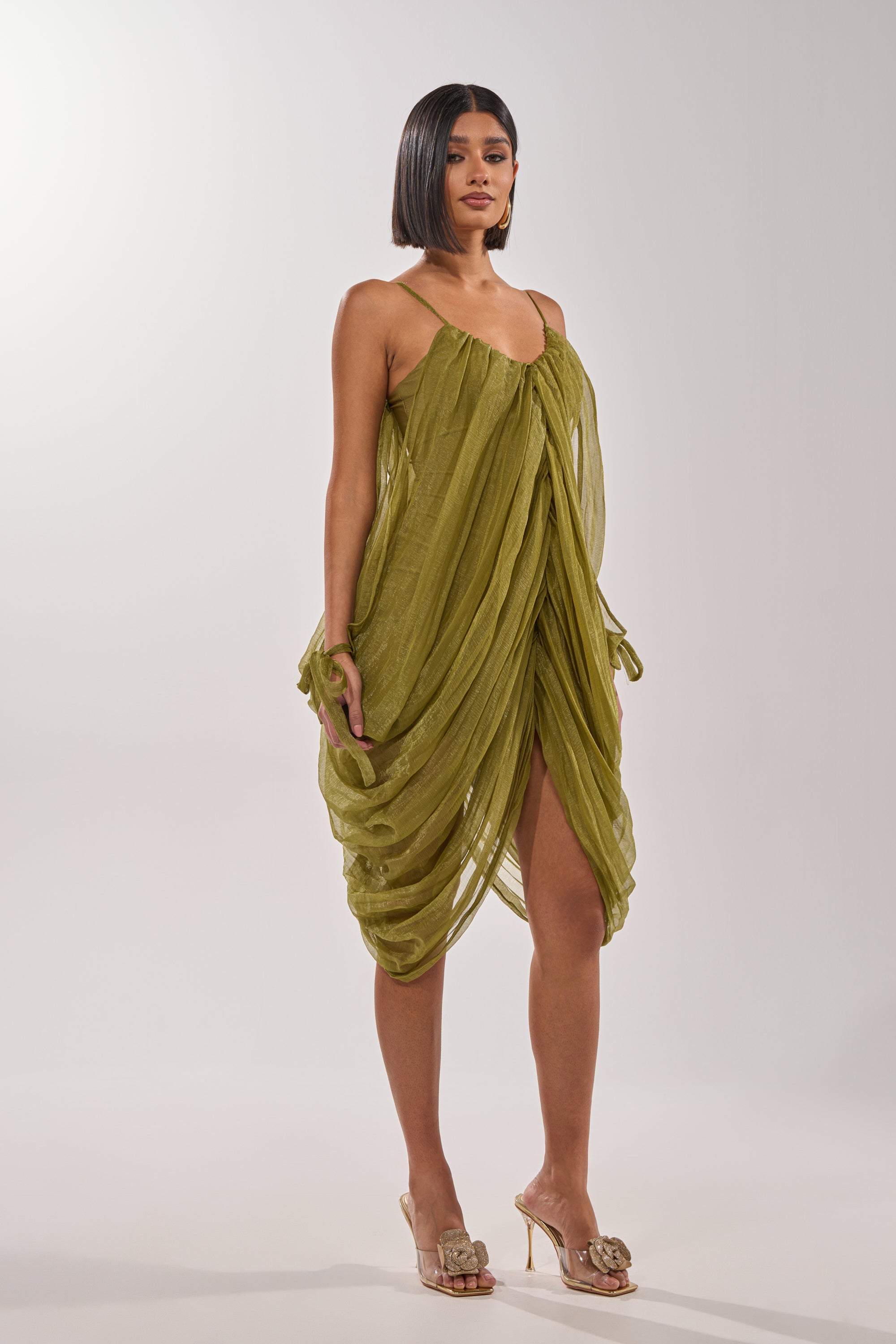 A woman poses against a plain light background in the VIVA LA VIDA MESH DRESS, a light olive draped style with thin straps, paired with gold heeled sandals. Her straight, chin-length hair completes the elegant look.