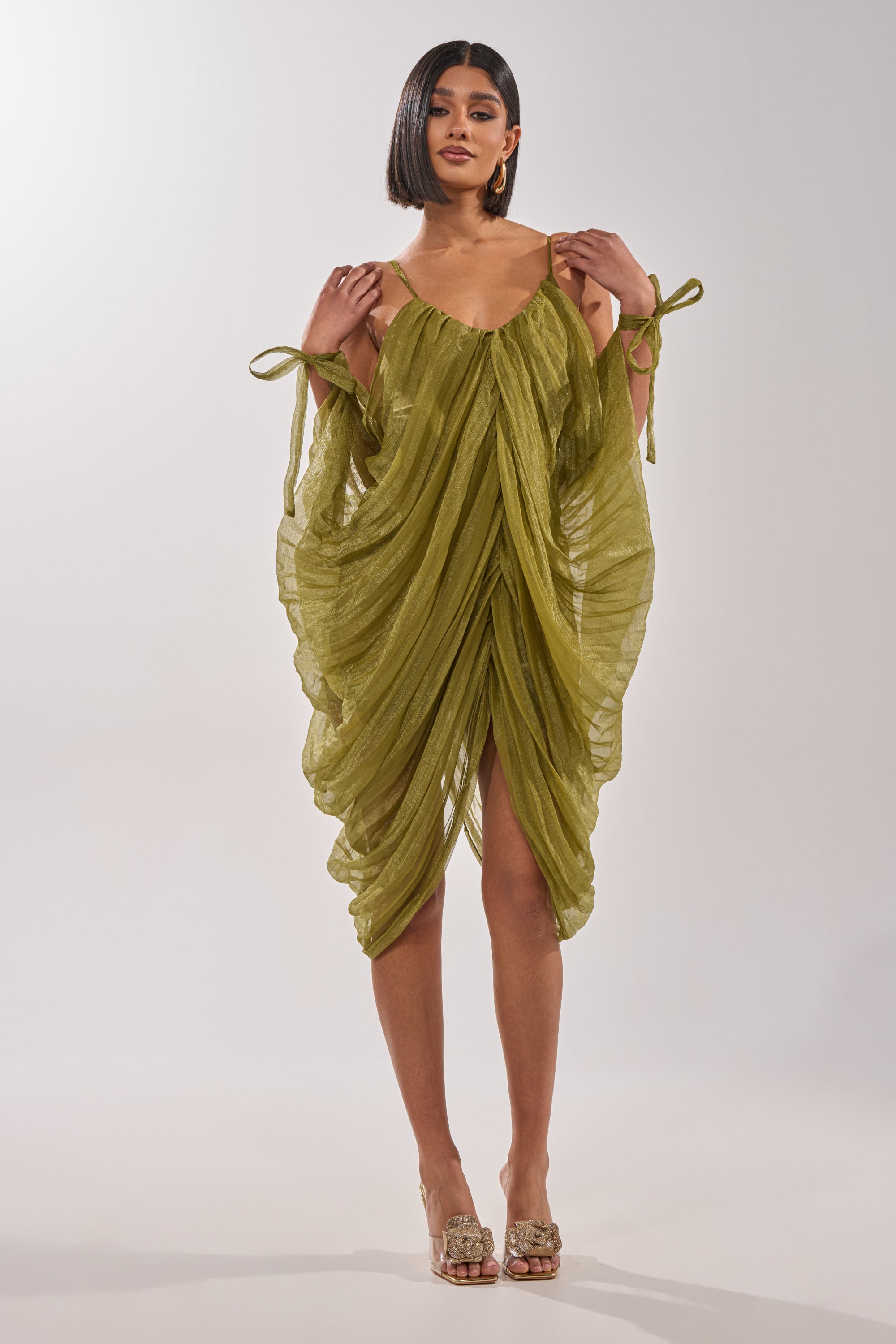 A woman with a sleek bob wears the VIVA LA VIDA MESH DRESS—an olive draped style with tied straps and open sleeves—paired with embellished open-toed heels. She stands against a plain white background. 2