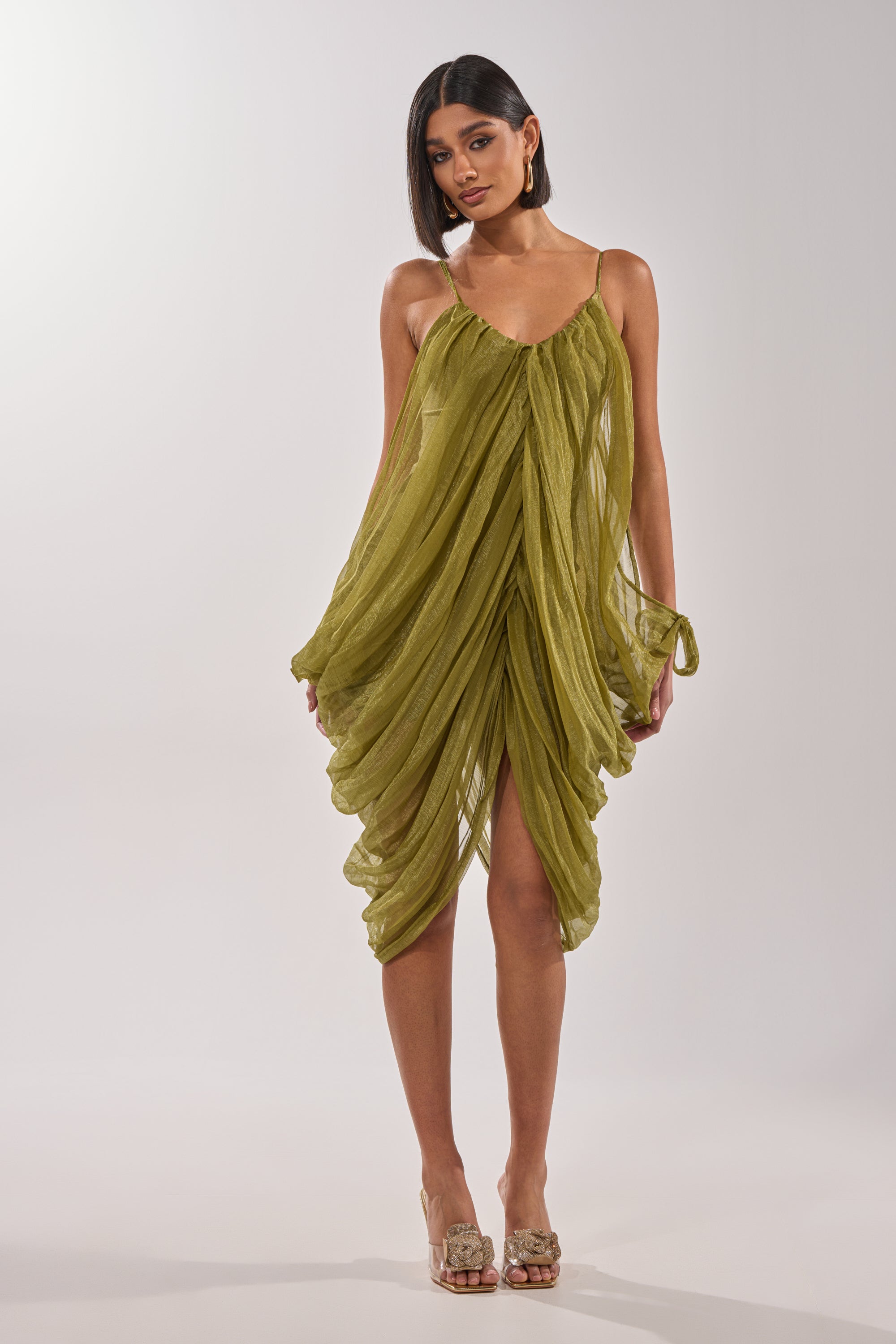 A woman wears the VIVA LA VIDA MESH DRESS, an olive midi dress with thin straps, a high-low hemline, and a light, airy mesh design. She styles it with shoulder-length straight hair, hoop earrings, and open-toe heels. 3