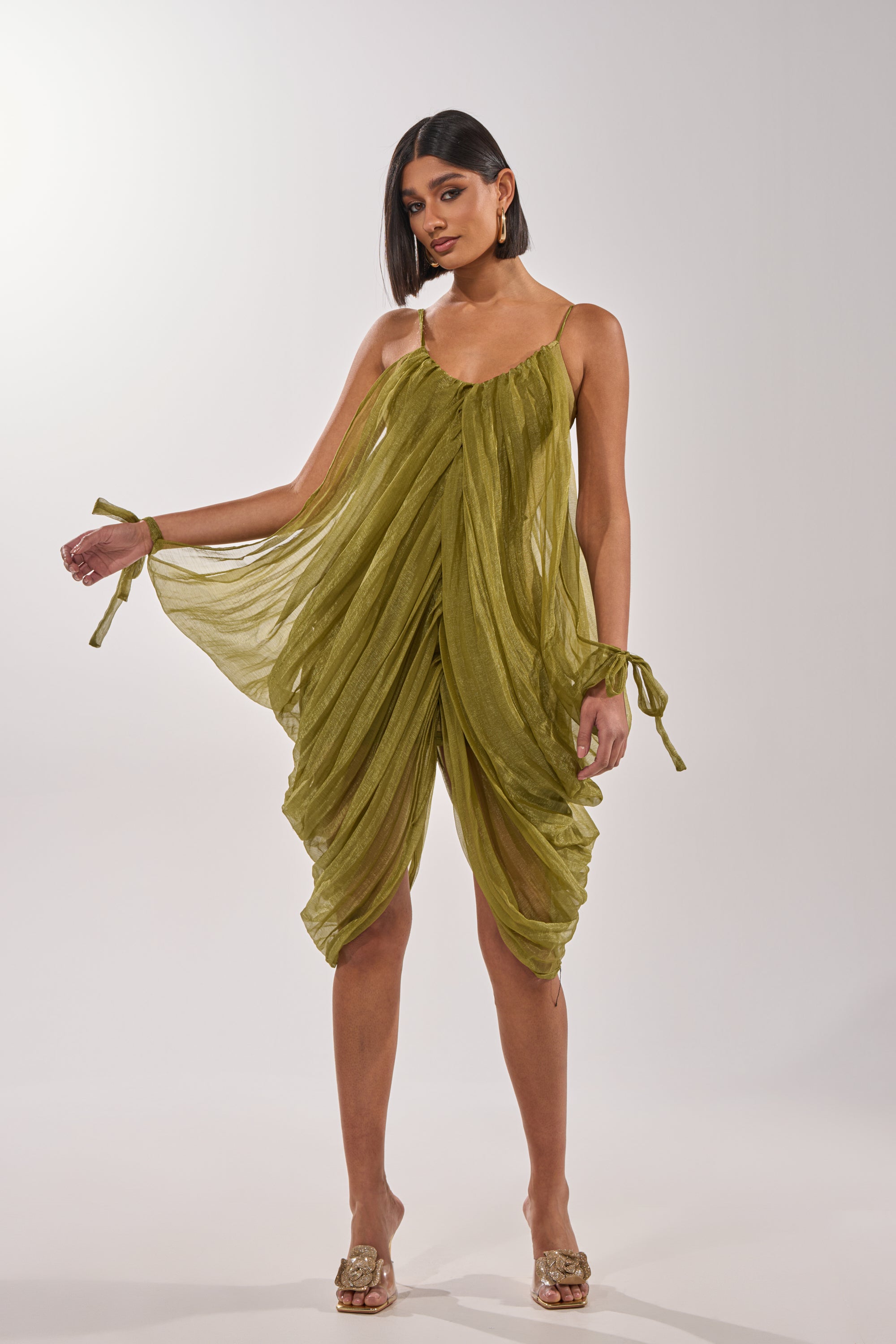 A woman wears the VIVA LA VIDA MESH DRESS in olive green with sheer mesh, draped details, thin straps, and wrist ties, styled with clear-heeled sandals against a plain white background. 0
