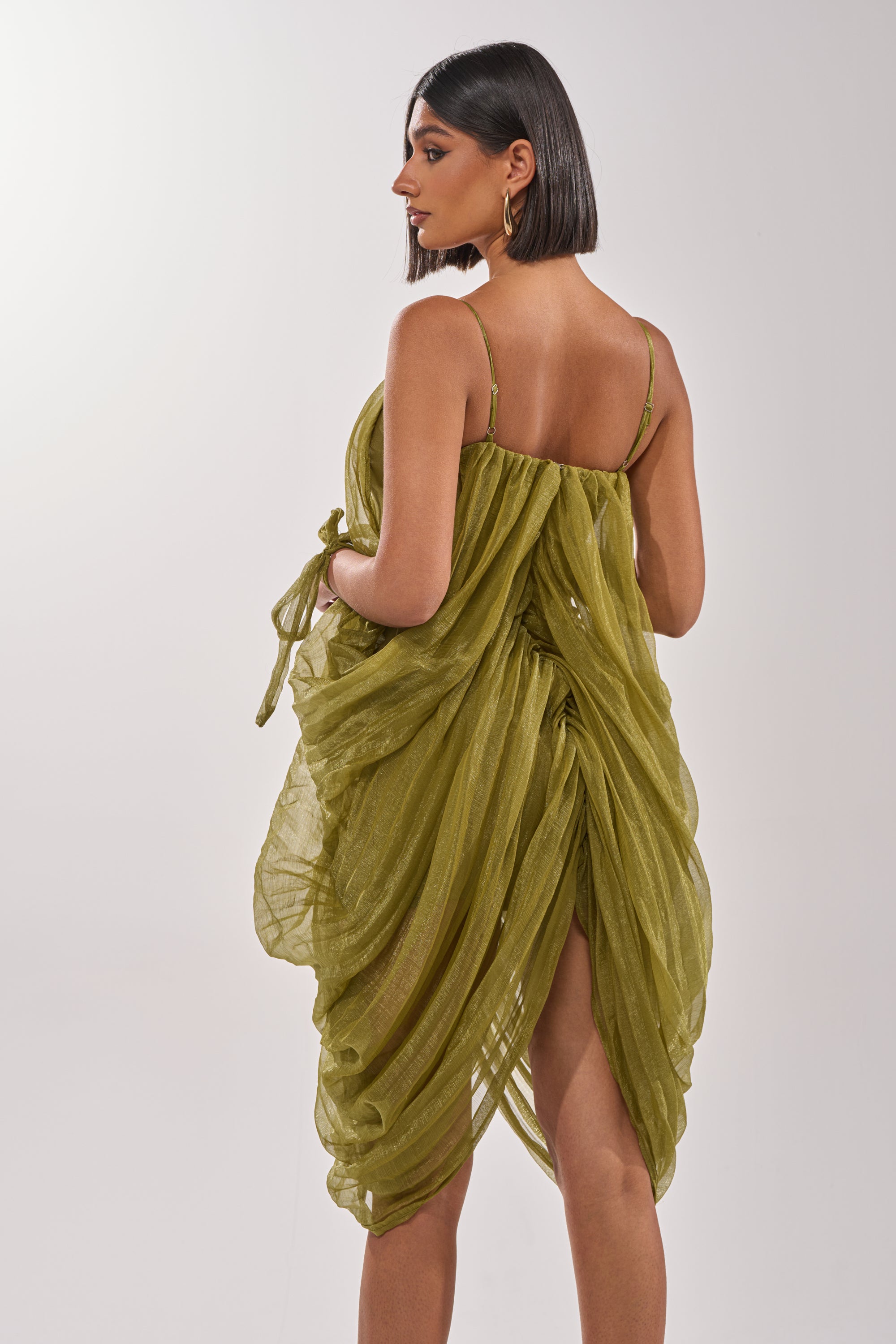 A woman with short dark hair wears the VIVA LA VIDA MESH DRESS, an olive draped style with thin straps, open back, and gathered details, standing against a plain light background. 5