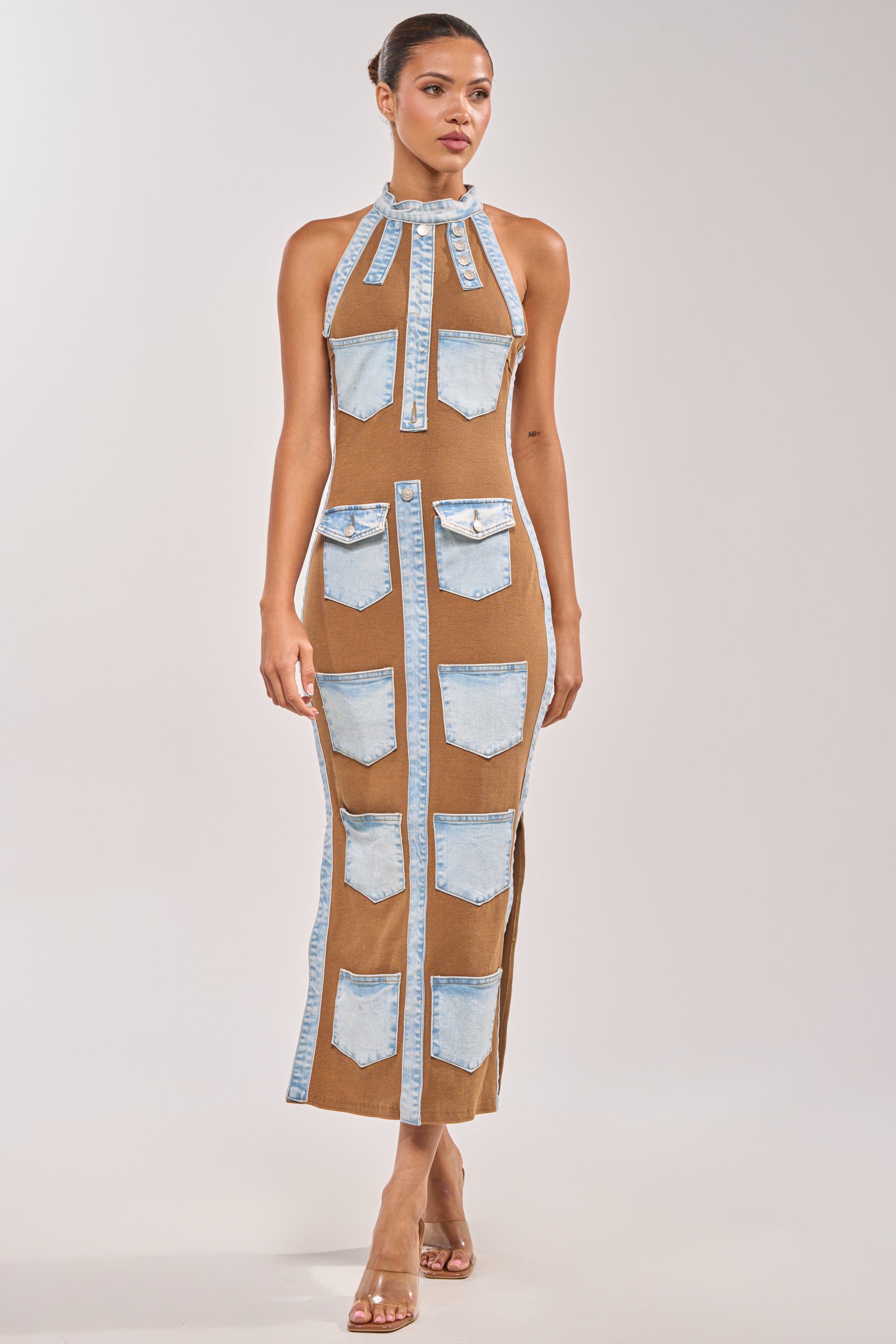 A woman wears the POCKET THEORY PATCHED UP MAXI, a brown bodycon maxi dress with denim pocket accents and a high neckline, styled with clear high-heeled sandals against a plain background. 0