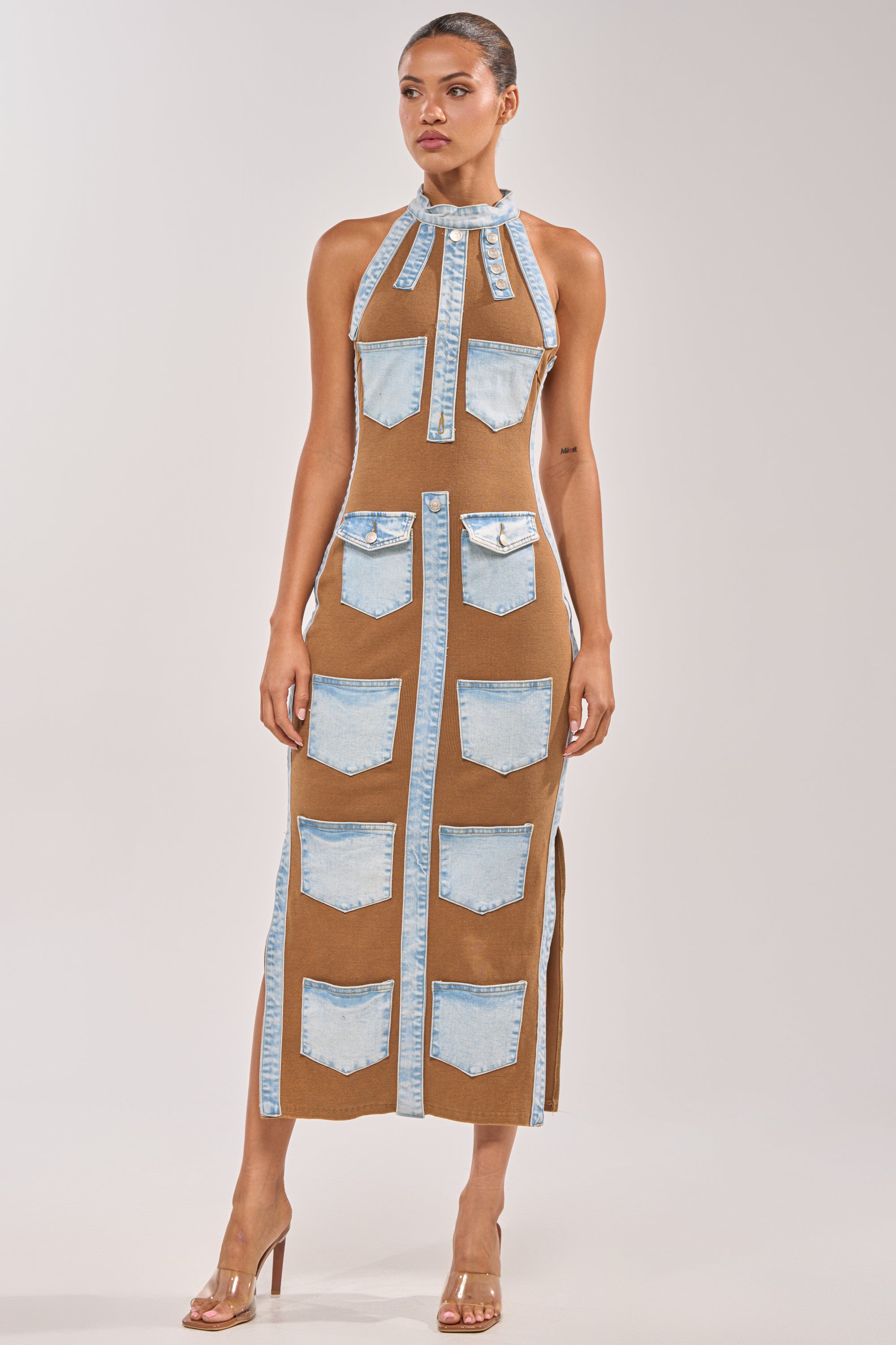 A woman wears the POCKET THEORY PATCHED UP MAXI, a brown sleeveless bodycon dress with a mockneck, denim pocket details, trim, and front ties. She finishes her streetwear-inspired look with clear high-heeled sandals against a plain background. 1