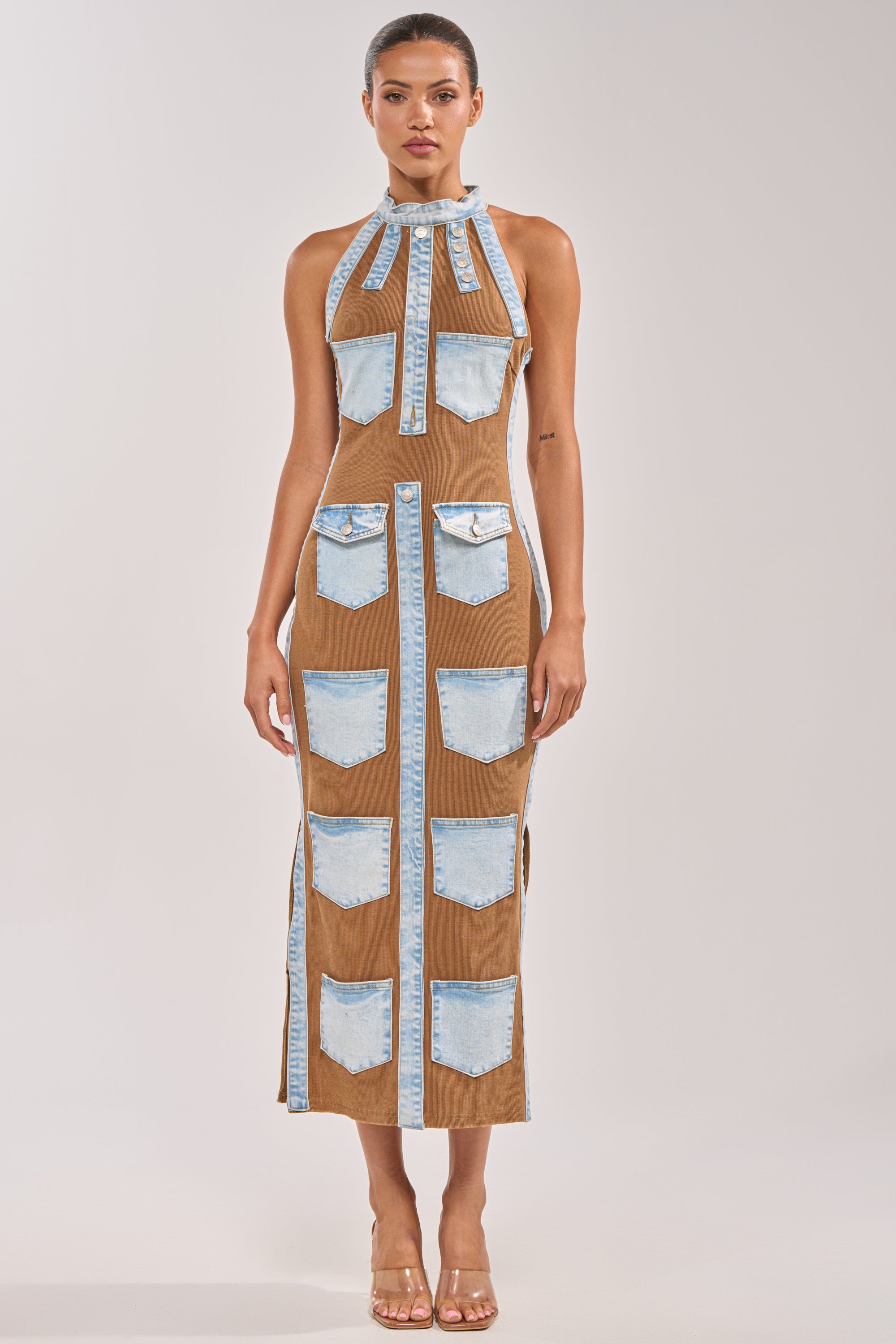 Wearing the POCKET THEORY PATCHED UP MAXI, a woman faces forward in a sleeveless, ankle-length brown dress featuring rows of denim pockets on the front. She pairs it with clear high-heeled shoes and has her hair pulled back. 4