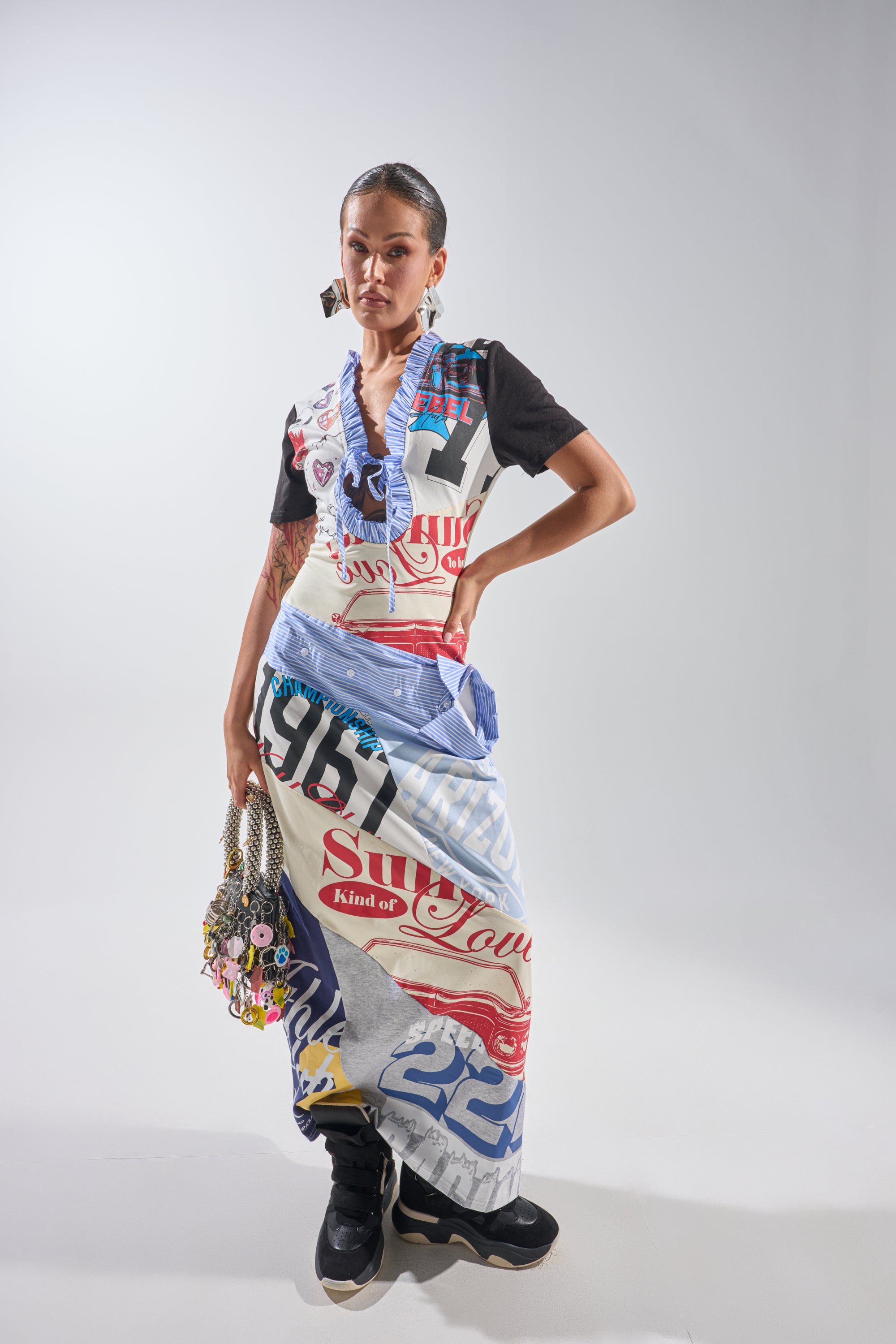 A woman poses confidently in the SOMETHING DIFFERENT MAXI DRESS, featuring bold graphic prints and text. She pairs it with chunky black sneakers, statement earrings, and a beaded bag, standing against a plain light gray background. 1