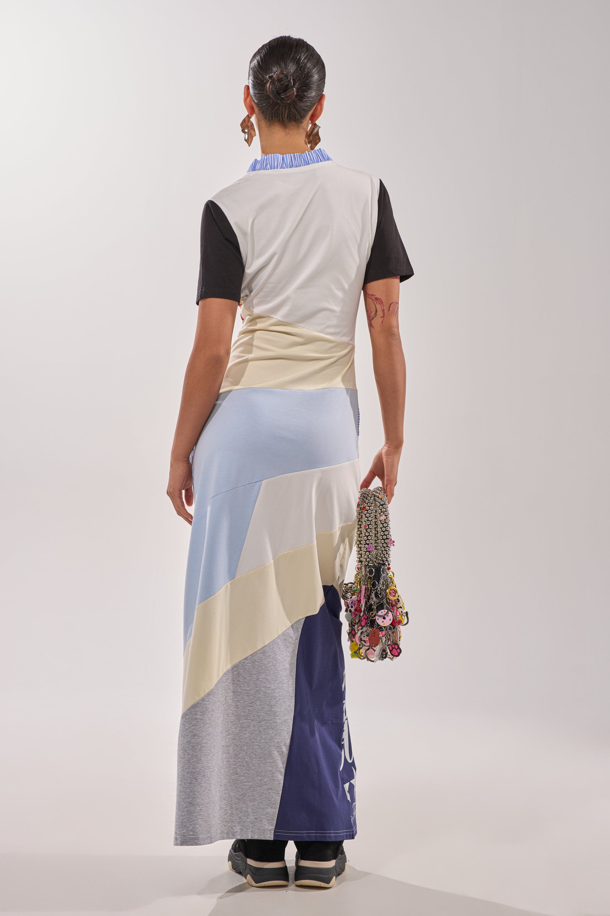 Wearing the SOMETHING DIFFERENT MAXI DRESS with short sleeves and geometric pastel panels, a person stands facing away, holding a floral bag in their right hand and their dark hair pulled back for an effortlessly chic look. 9