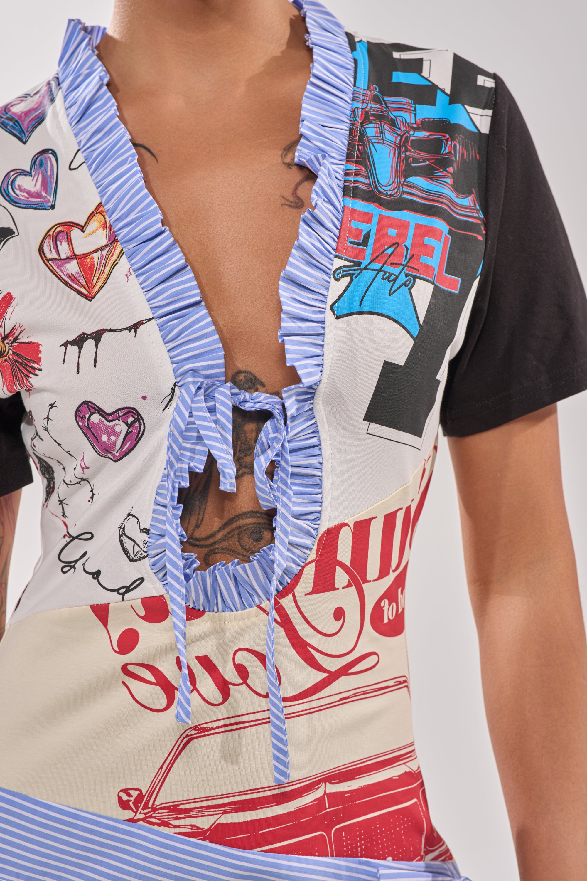 Someone wears the SOMETHING DIFFERENT MAXI DRESS—a colorful, short sleeve graphic maxi with a deep V-neckline trimmed in blue ruffles and heart and car prints. The dress ties at the neckline, revealing a partially visible chest tattoo. 10