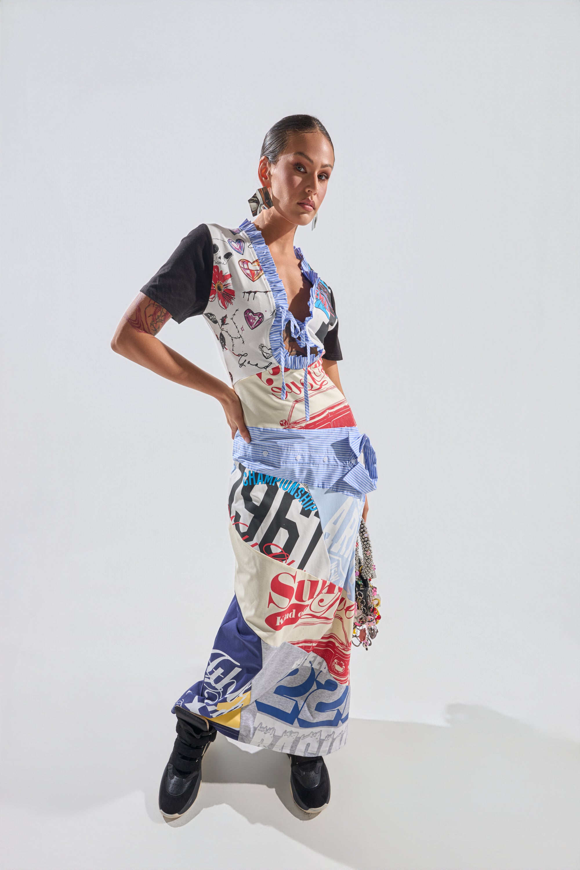 A person poses confidently in the SOMETHING DIFFERENT MAXI DRESS, a colorful collage print made from branded and graphic fabrics with bold logos. They wear black boots, statement earrings, and have slicked-back hair against a plain white background. 2