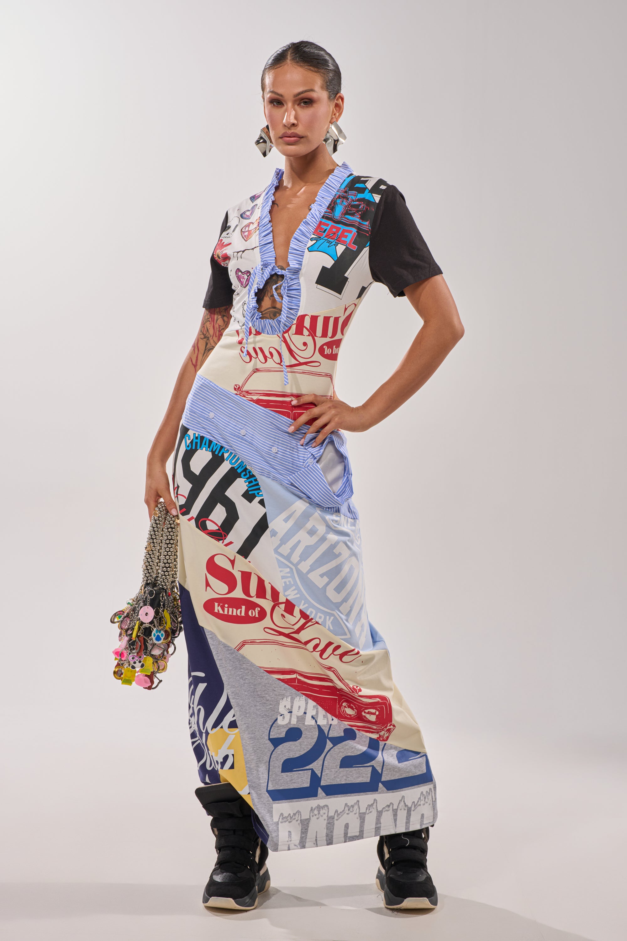 A woman stands confidently in the SOMETHING DIFFERENT MAXI DRESS, featuring a vibrant collage print with mixed designs and text. She carries a chain strap bag with keychains and pairs the look with black sneakers against a plain white background.
