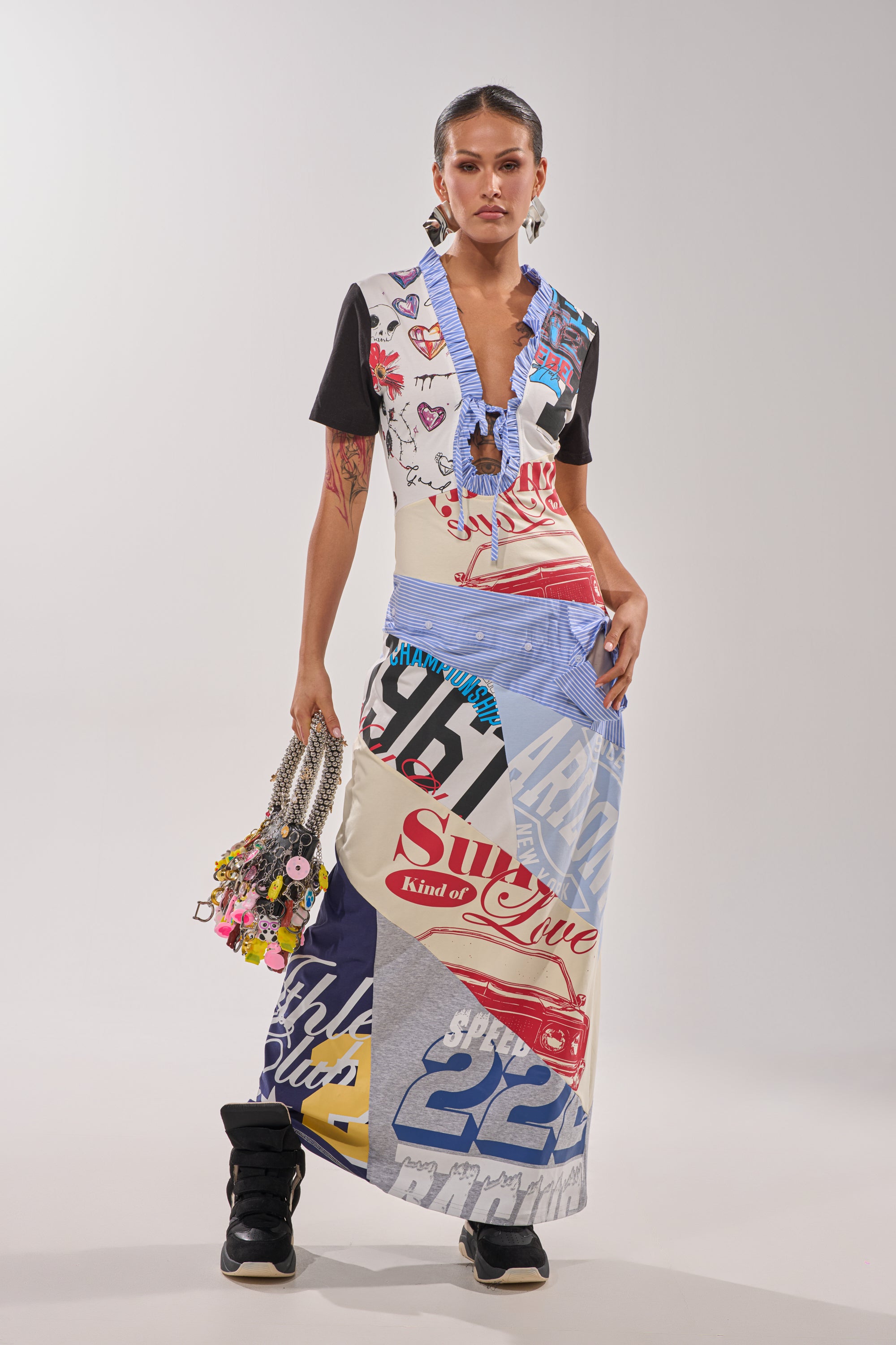 A woman poses in the SOMETHING DIFFERENT MAXI DRESS, a colorful eclectic outfit with bold graphics, paired with chunky sneakers, statement earrings, and a beaded handbag adorned with charms. 4