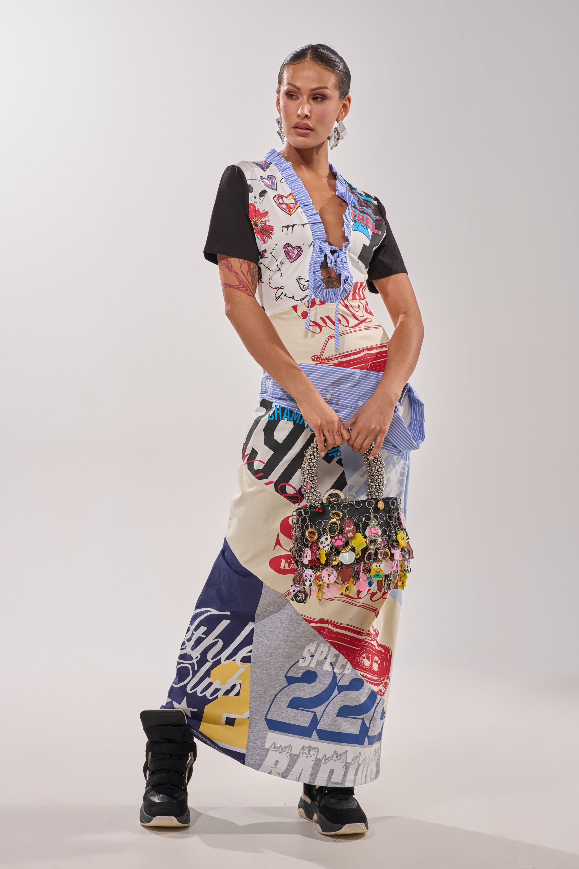 A woman stands confidently in the SOMETHING DIFFERENT MAXI DRESS—a collage print piece with bold graphics—paired with black sneakers and a handbag adorned with keychains, her sleek hair styled back as she poses against a plain background. 5