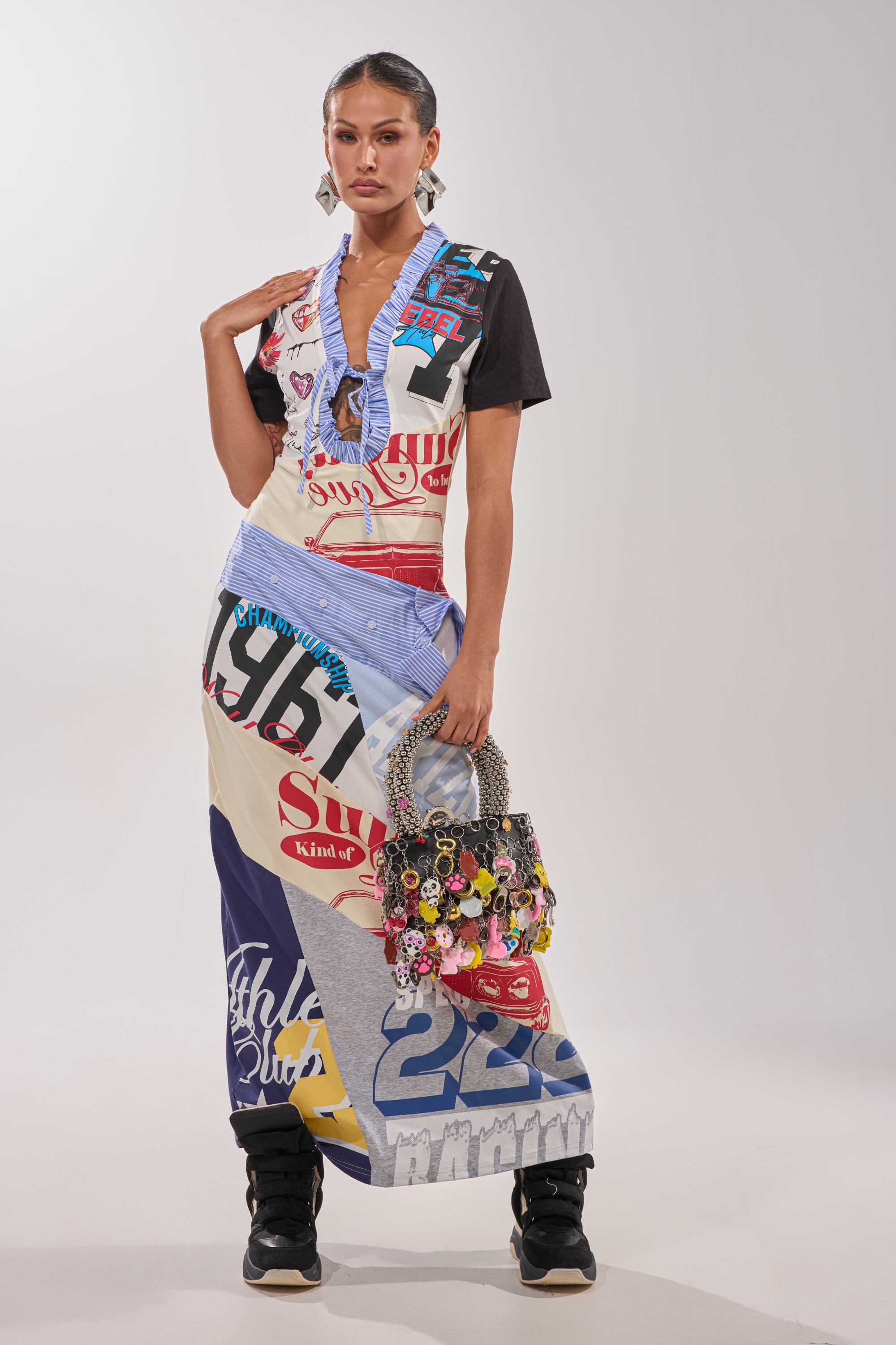 A woman stands against a plain background wearing the SOMETHING DIFFERENT MAXI DRESS over a black t-shirt, paired with black sneakers and holding a beaded handbag adorned with assorted charms. 6