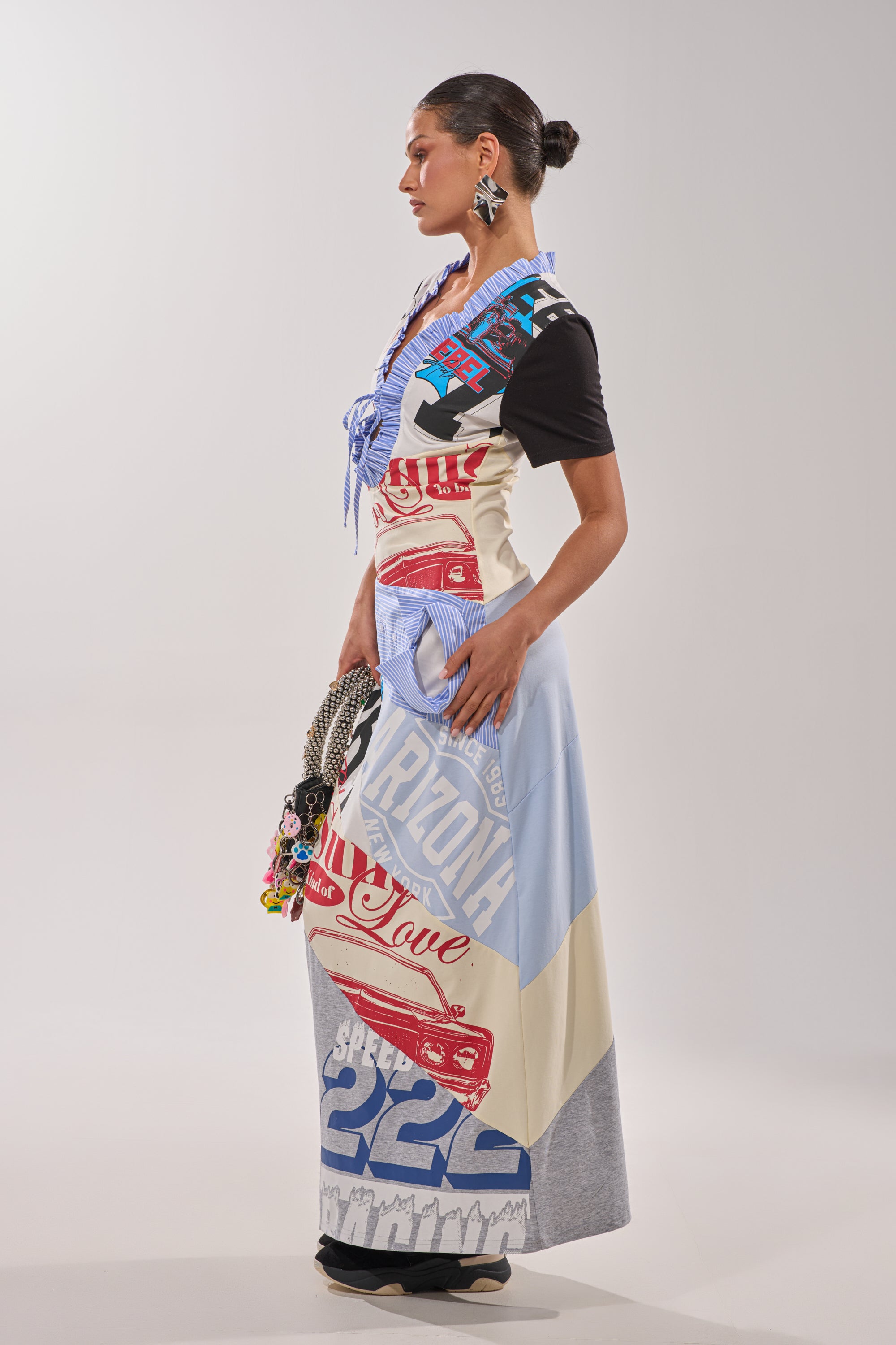 A woman stands sideways in the SOMETHING DIFFERENT MAXI DRESS, a graphic collage-print maxi dress in blue, red, and white with bold car and text motifs. She finishes her striking look with slicked-back hair, statement earrings, and a beaded handbag. 7