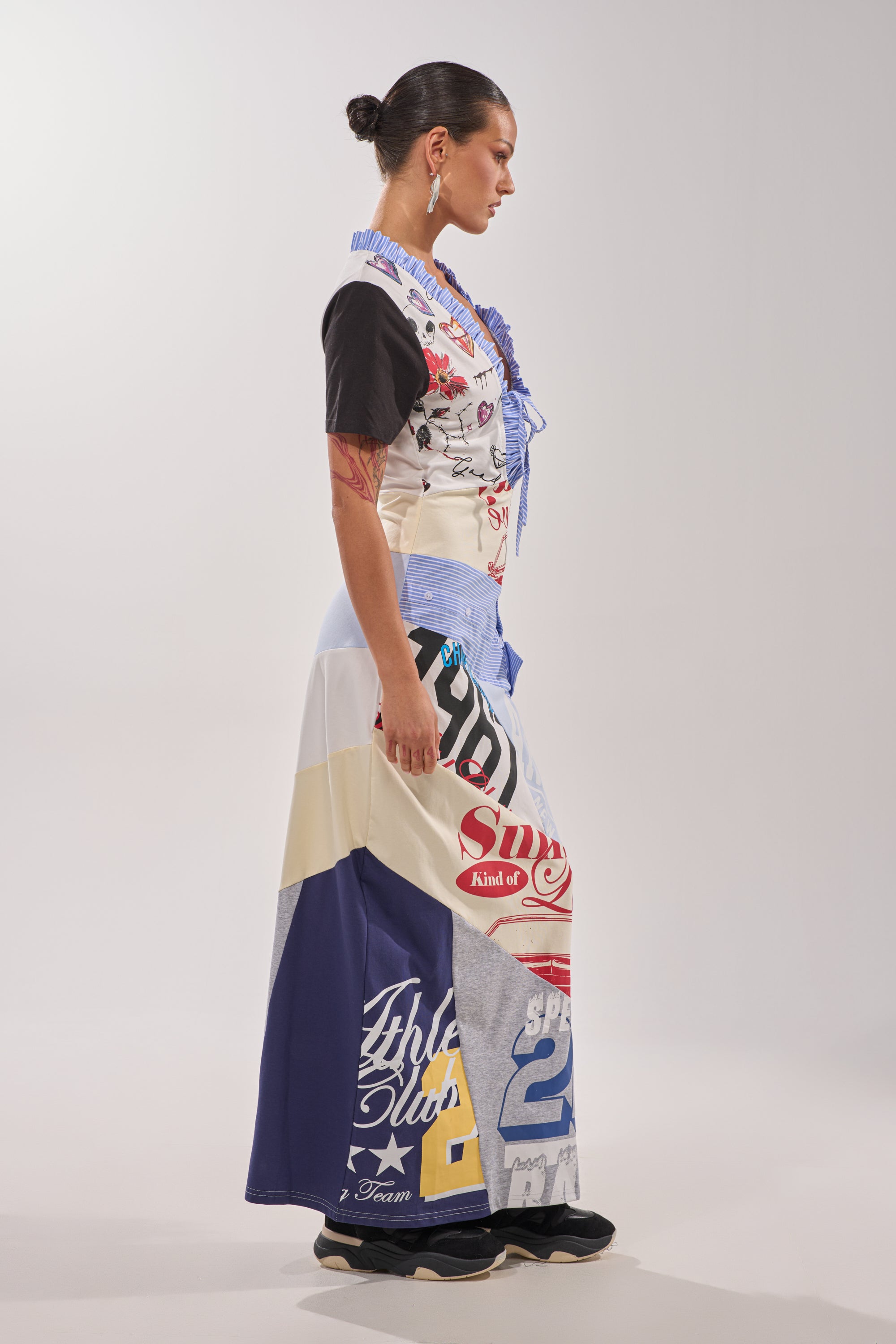 A woman stands in profile against a plain light background, wearing the SOMETHING DIFFERENT MAXI DRESS—a vibrant collage-print style made from graphic t-shirts. She has slicked-back hair, hoop earrings, and black shoes. 8